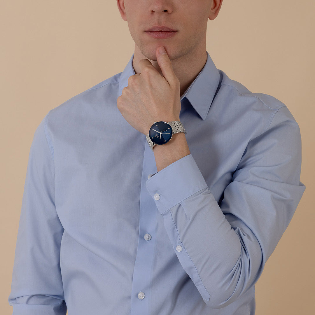A person in a light blue button-up shirt poses with one hand touching their chin, wearing the RADO Florence Automatic 39mm Watch by RADO, which features a dark face and durable sapphire crystal. The background is plain beige.
