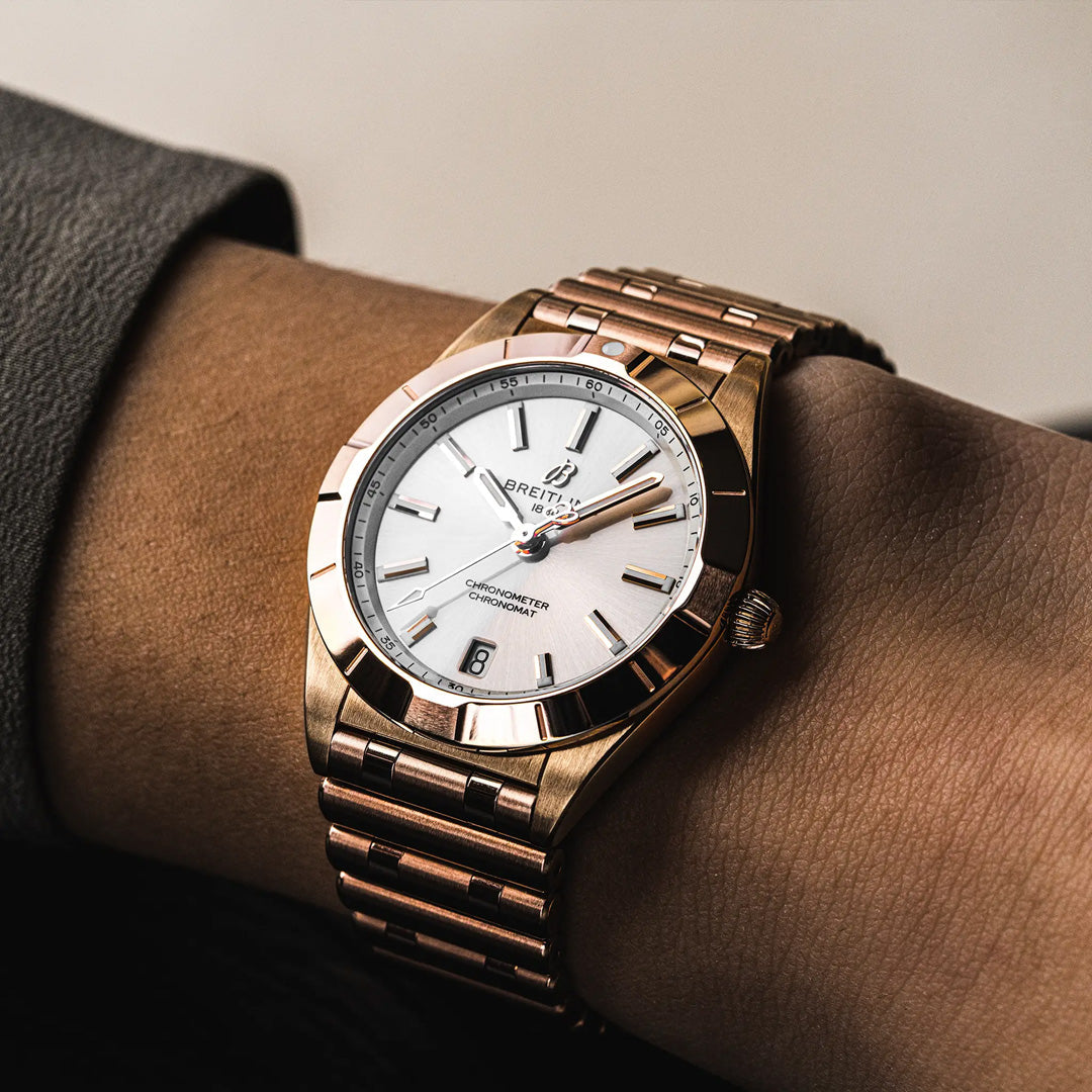 A close-up of the BREITLING Chronomat 32 by Breitling shows its gold case, white dial, and metal band elegantly worn on a wrist. This timepiece features a detailed diamond-set bezel, striking hour markers, and displays both the time and date.