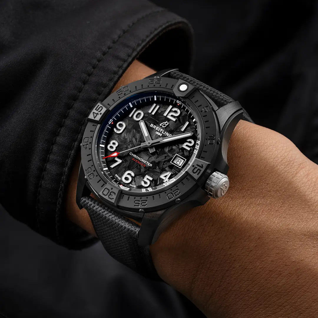 A person wears a BREITLING Avenger Automatic 42 Night Mission wristwatch, boasting a textured black strap and bold white numbers on a black dial. The aviation-inspired design includes three hands and a date display. Their wrist, in a dark jacket, contrasts with the neutral background.