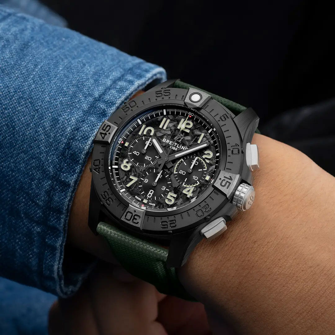 A close-up of a person wearing the Breitling Avenger B01 Chronograph 46 Night Mission, featuring a black face with large white numerals, multiple dials, and a green strap, styled over a blue denim sleeve.