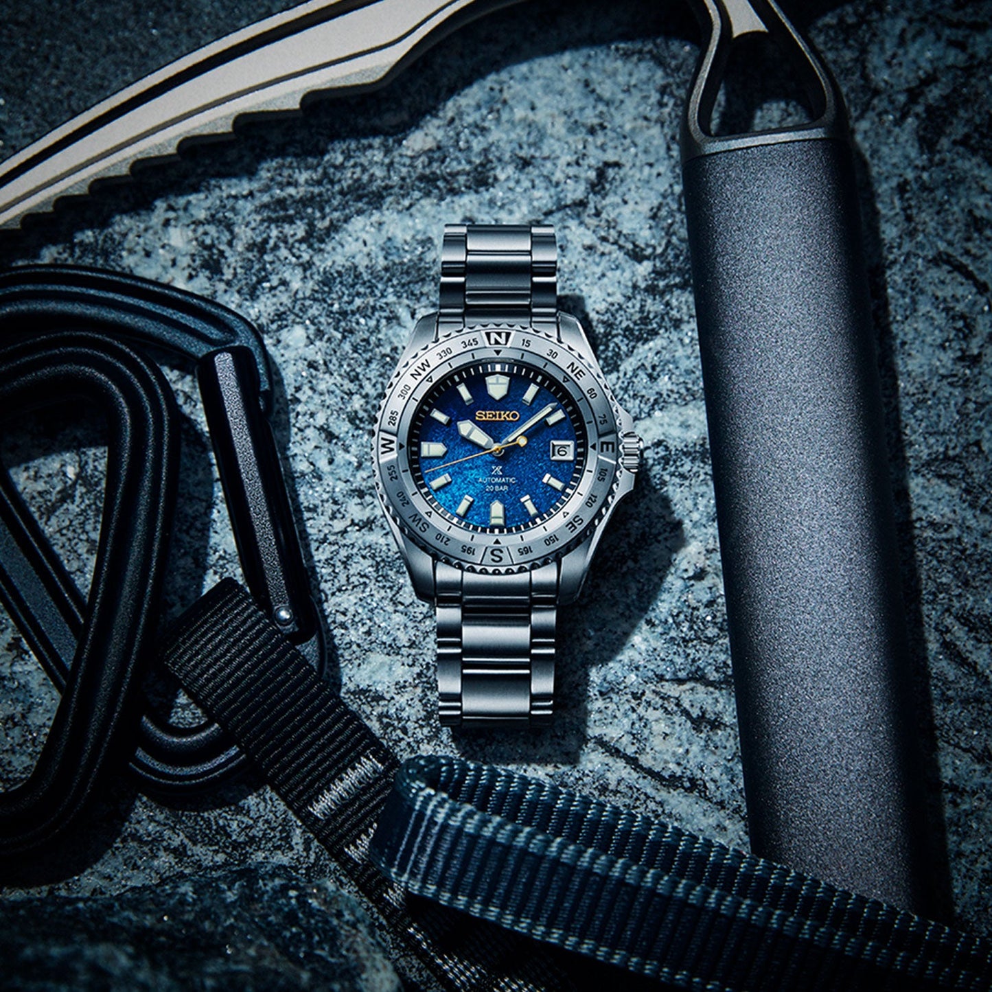 The Seiko Prospex Landmaster 30th Anniversary Limited Edition Automatic with Manual Winding 42mm watch, featuring a striking blue dial and a sleek silver band, is artfully displayed on a rugged rocky surface. It is surrounded by climbing gear such as ropes and a metal tool, perfectly capturing an adventurous theme that celebrates three decades of exploration excellence.