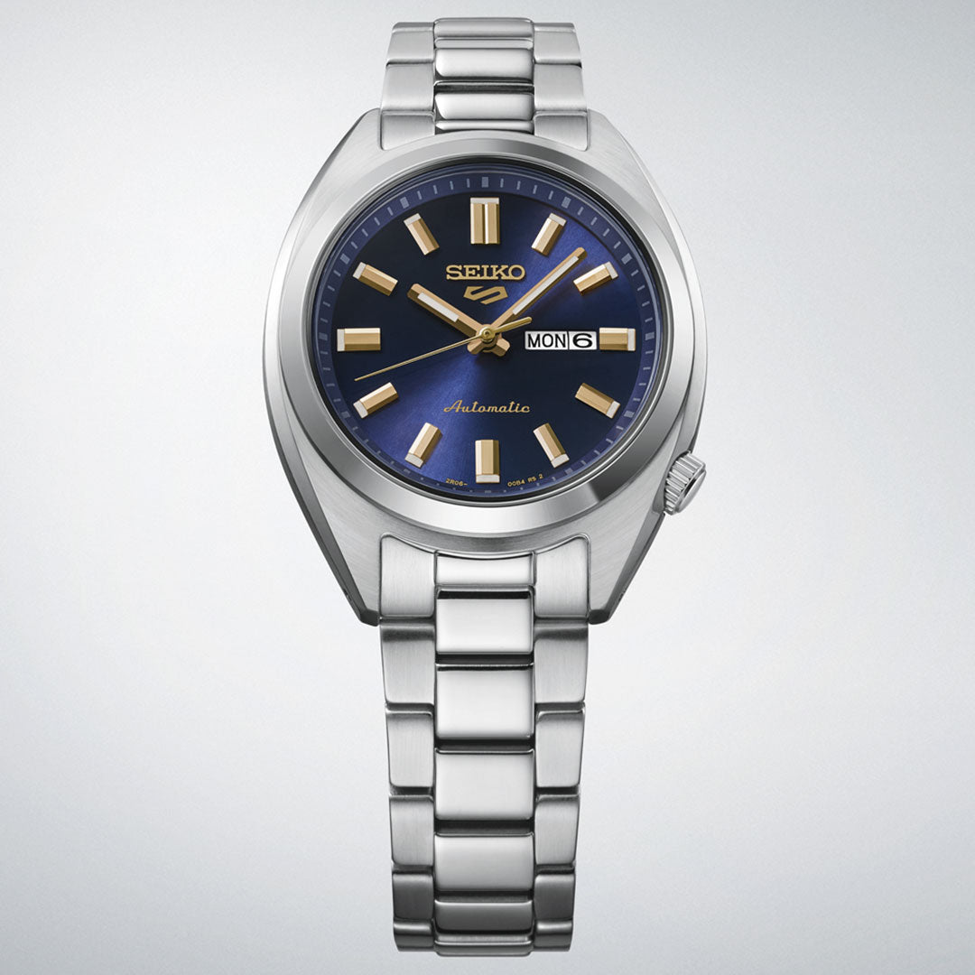 The Seiko 5 Sports SNXS Series Automatic 32mm Watch features a blue and gold dial, gold hour markers, silver bracelet, and a day-date display at 3 o’clock. Brand: Seiko. Set against a light gradient background.