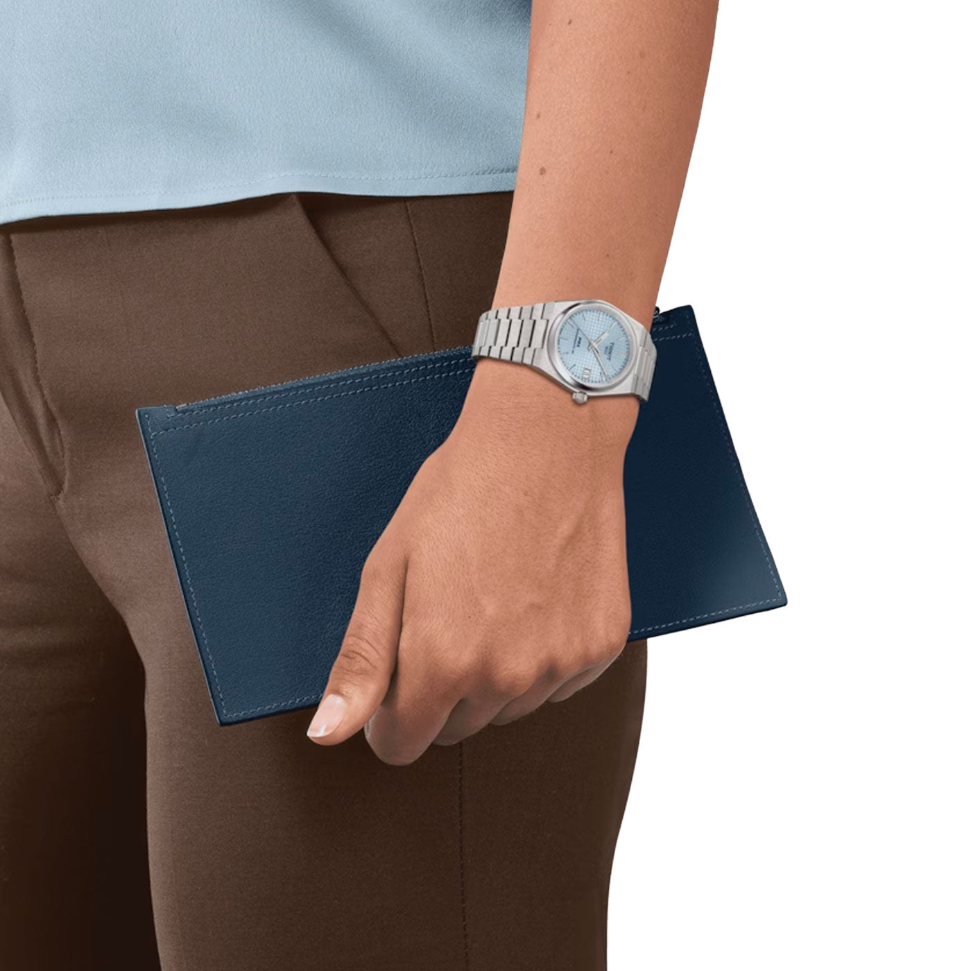 A person dressed in a light blue shirt and brown pants is holding a dark blue wallet. On their wrist, they are wearing the Tissot PRX Powermatic 80 Automatic 35mm Watch, featuring a silver stainless steel case. The background is plain white.