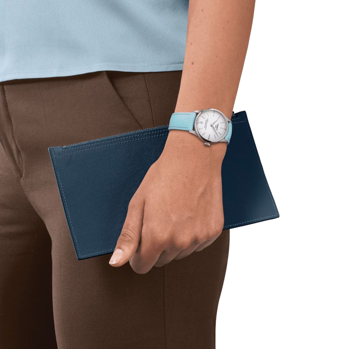 A person in a light blue shirt and watch, showcasing the precision of the Tissot Chemin Des Tourelles Powermatic 80 Automatic 34mm Watch by Tissot, holds a dark blue clutch. They are dressed in brown pants. The image focuses on the arm and hand, emphasizing the clutch and watch.