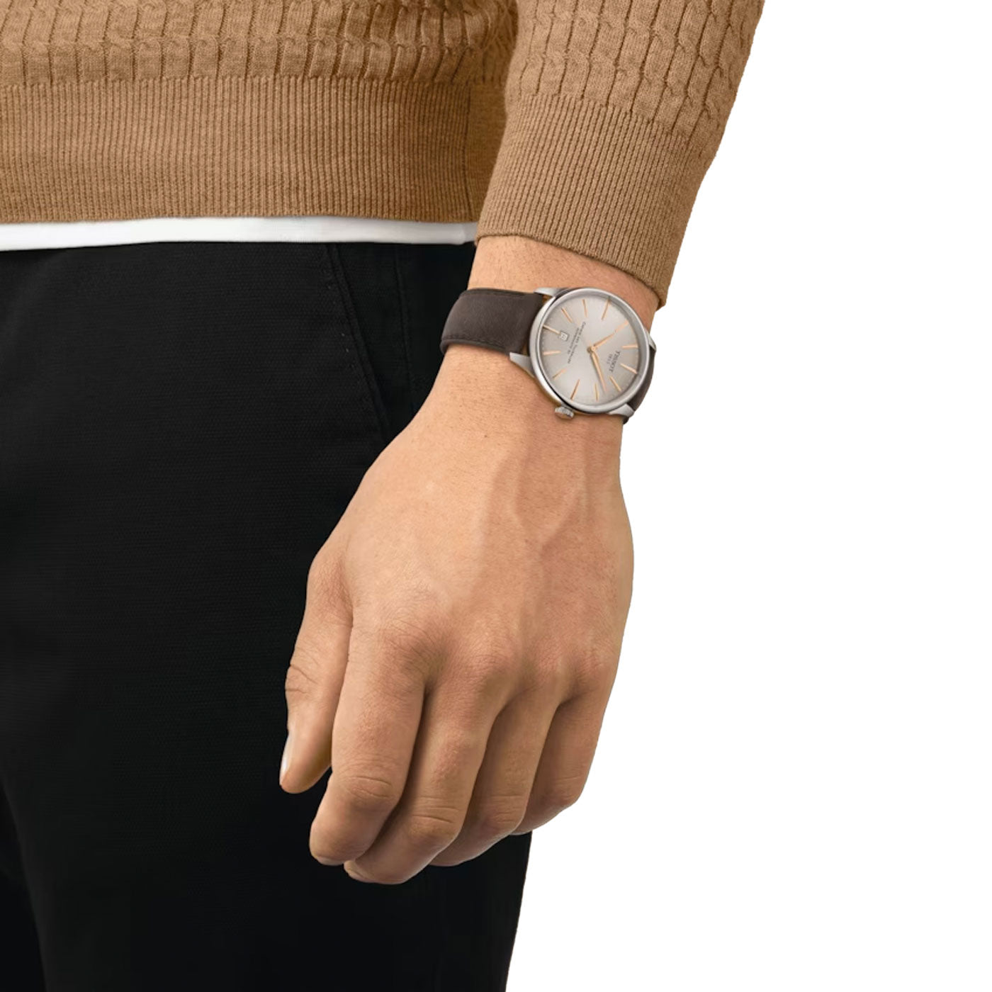 A person wearing a brown sweater and black pants displays the Tissot Chemin Des Tourelles Powermatic 80 Automatic 42mm Watch. This elegantly designed Tissot timepiece, featuring a brown leather strap and a silver watch face with simple hour markers, showcases refined precision thanks to its Powermatic 80 movement.