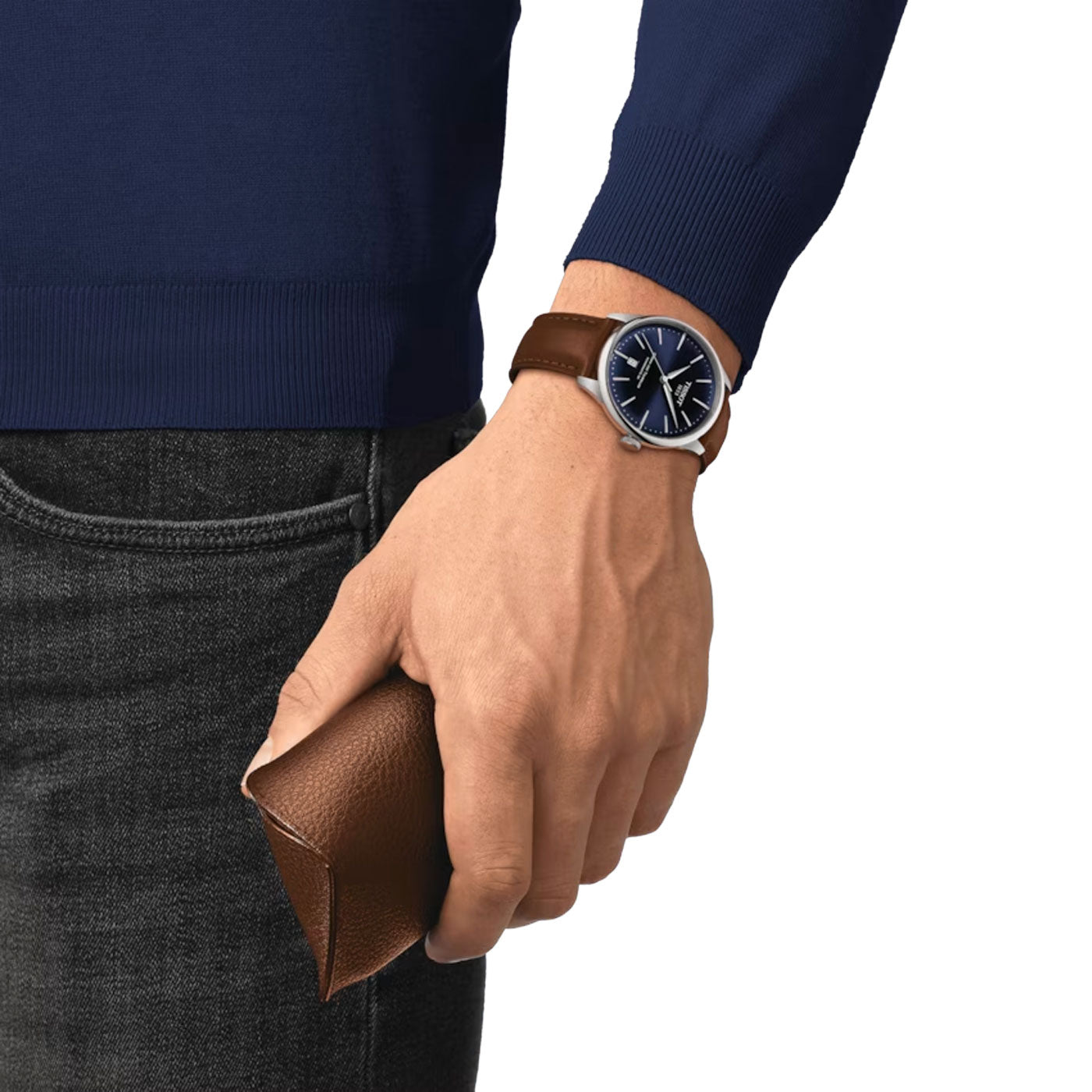 A person donning a dark blue long-sleeve shirt and jeans holds a brown leather clutch. On their wrist is the Tissot Chemin Des Tourelles Powermatic 80 Automatic 42mm Watch by Tissot, featuring an interchangeable quick-release bracelet with a dark blue face and matching brown strap, possibly concealing the precise Powermatic 80 movement within.