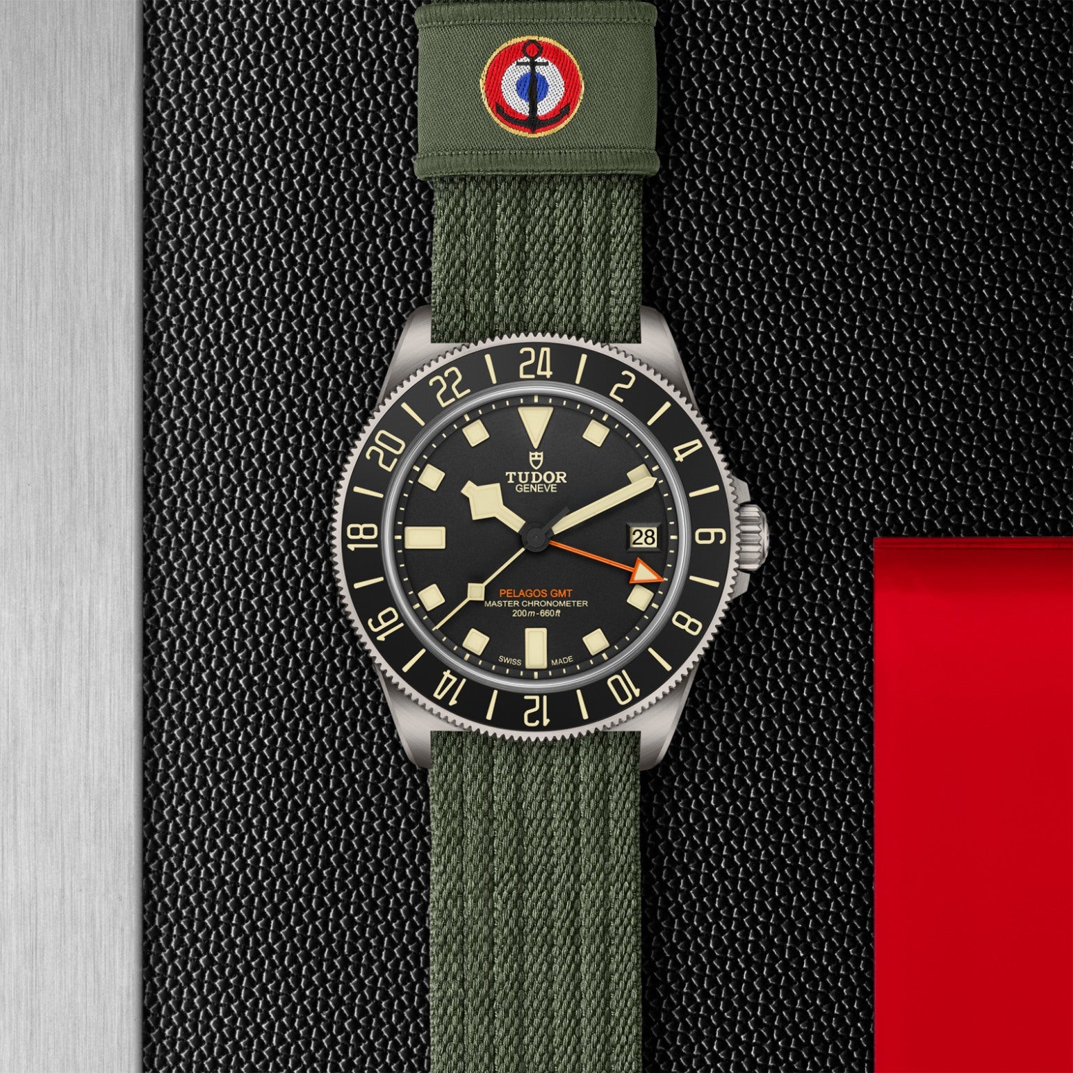 The TUDOR Pelagos FXD GMT wristwatch by TUDOR features a black dial, beige markers, and a green fabric strap with red and blue embroidery, inspired by French Naval Aviation, shown on a textured black background with metallic and red details.