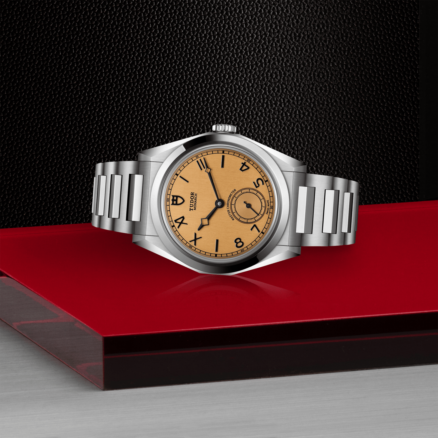 The TUDOR Monarch by TUDOR features a silver metallic band, a tan dial with black hour markers, and a small seconds subdial. It rests on a glossy red surface against a textured black background.