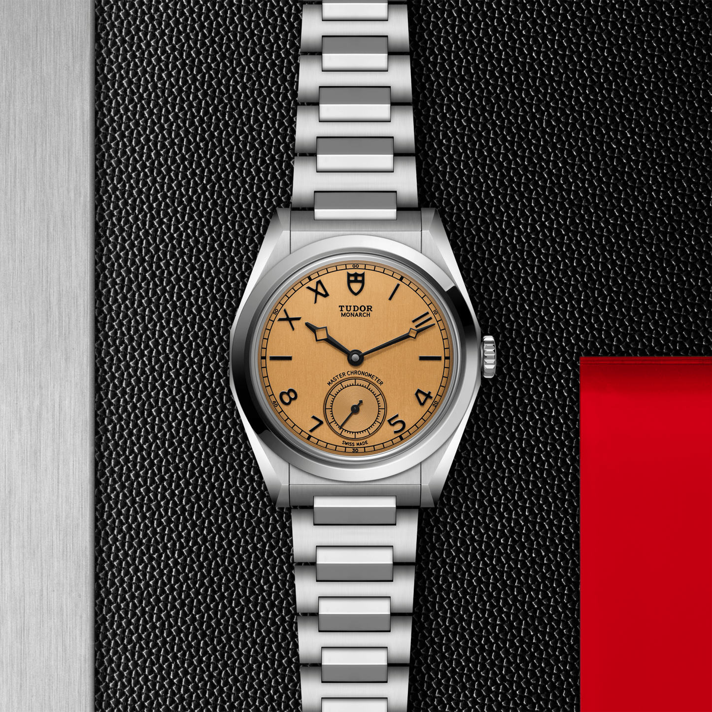 A TUDOR Monarch luxury watch with a metal bracelet, silver case, champagne dial, black numerals, and subdial is displayed on a textured black and red background with a brushed metal edge.