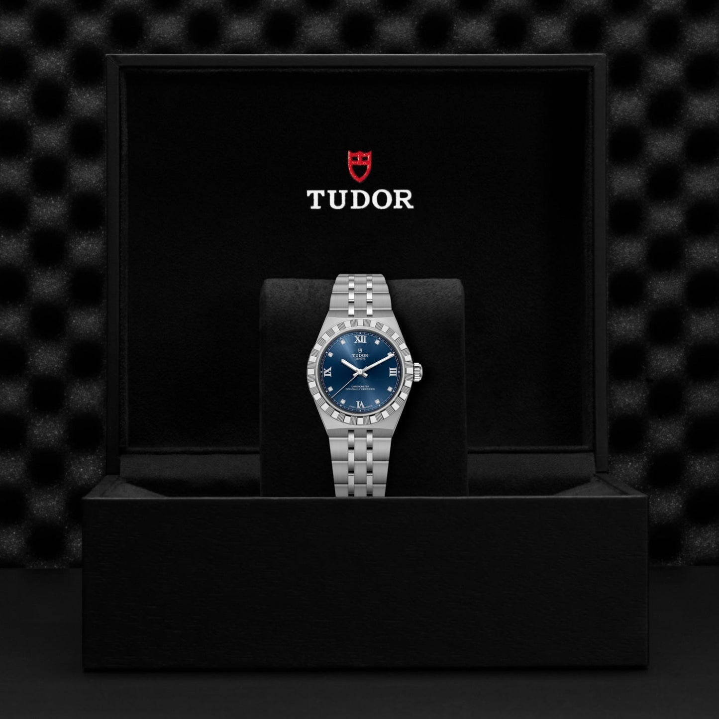 A TUDOR Royal wristwatch with a blue dial is presented in an open black box featuring the TUDOR logo and shield emblem on the lid, set against a textured black background that emphasizes TUDOR’s versatile sport-chic automatic watches.