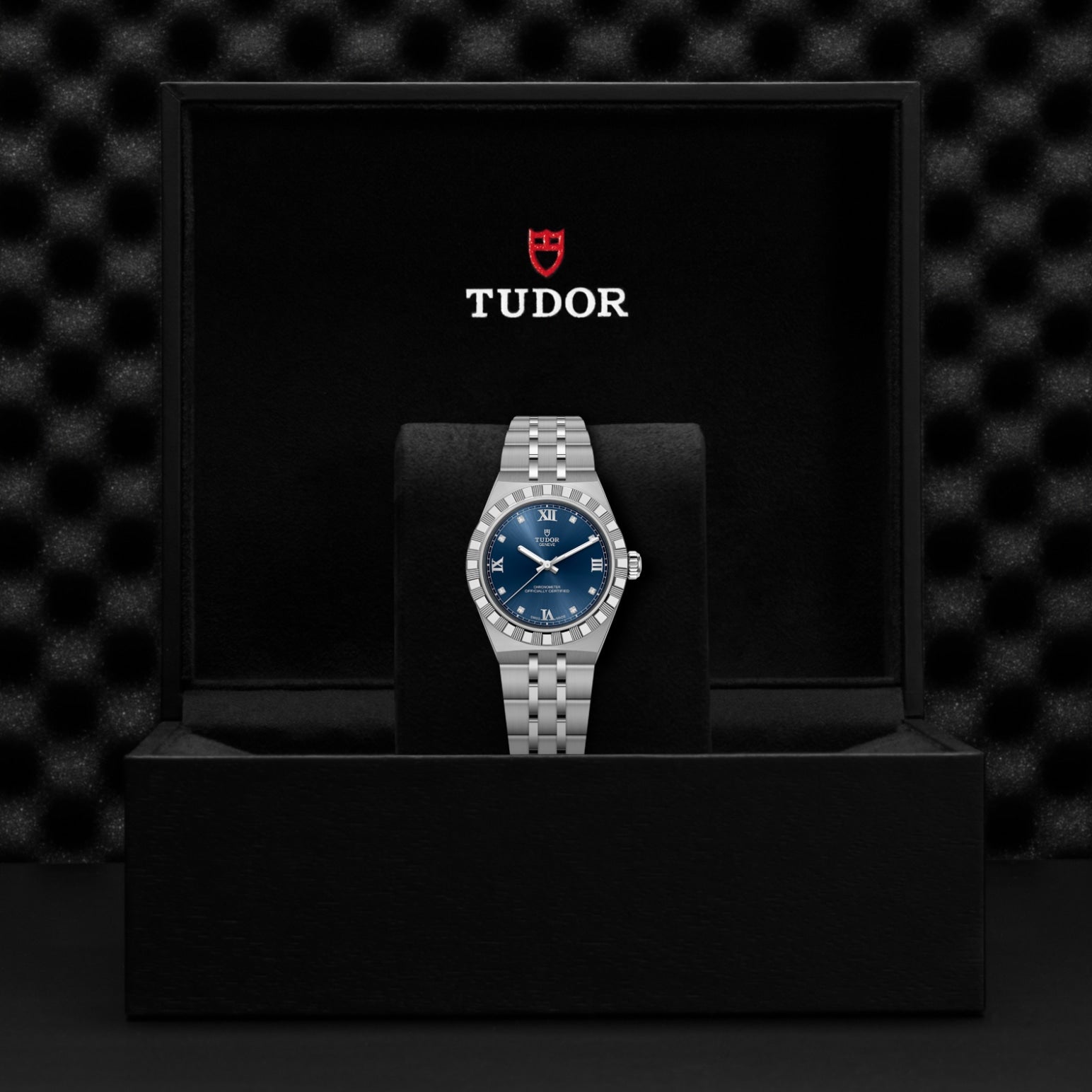 A TUDOR Royal wristwatch with a blue dial is presented in an open black box featuring the TUDOR logo and shield emblem on the lid, set against a textured black background that emphasizes TUDOR’s versatile sport-chic automatic watches.