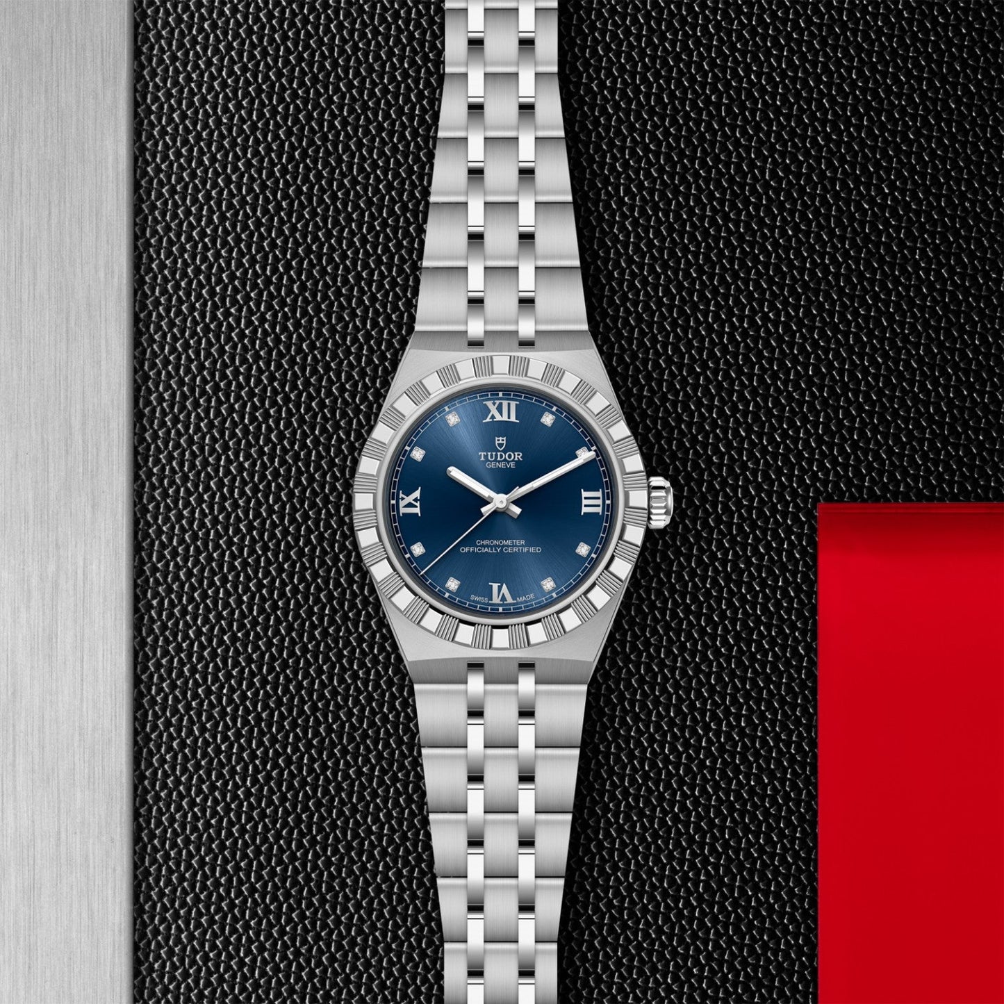 The TUDOR Royal by TUDOR, featuring a stainless steel case, blue dial with Roman numerals, and five-link bracelet, is showcased on a textured black background with silver and red accents for a sport-chic look.