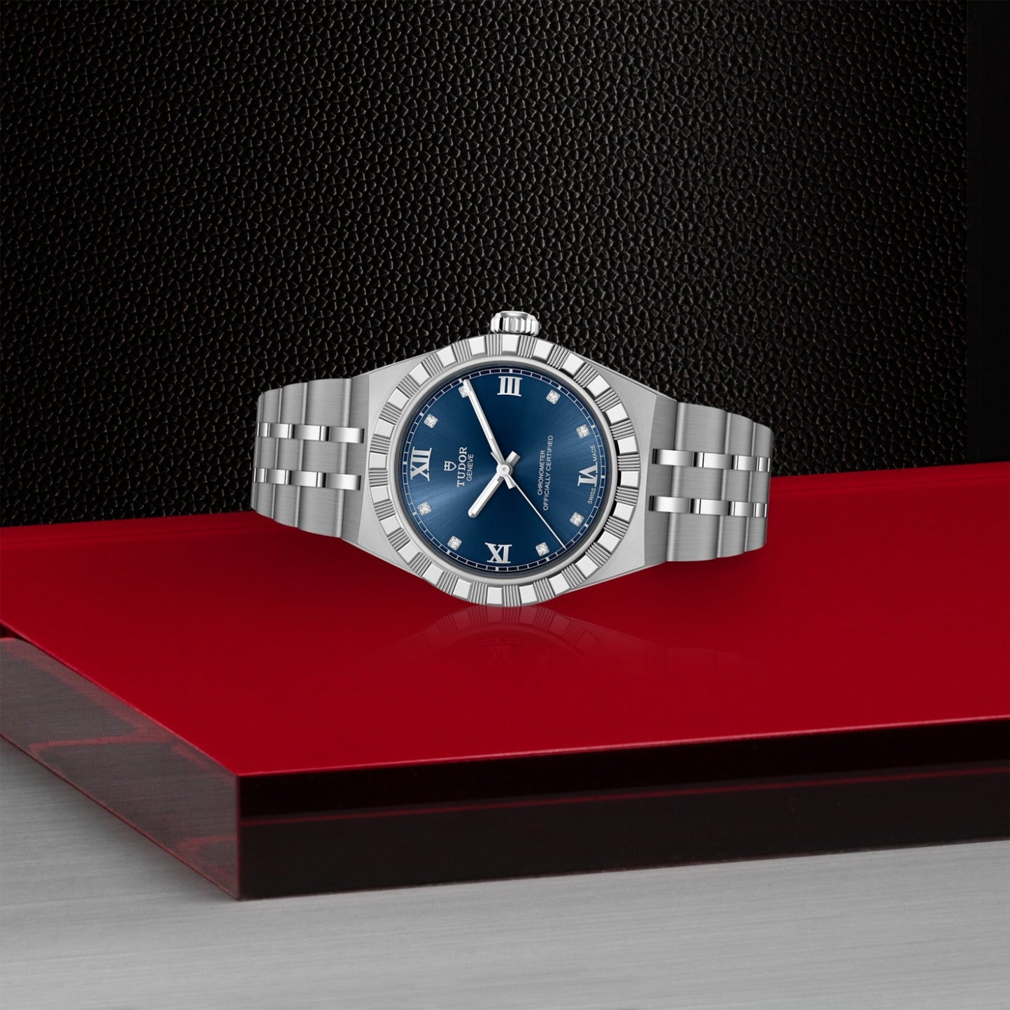 The TUDOR Royal by TUDOR, featuring a silver case, blue dial, and Roman numerals, rests on a red surface against a black textured background—an elegant symbol of automatic sport-chic style.