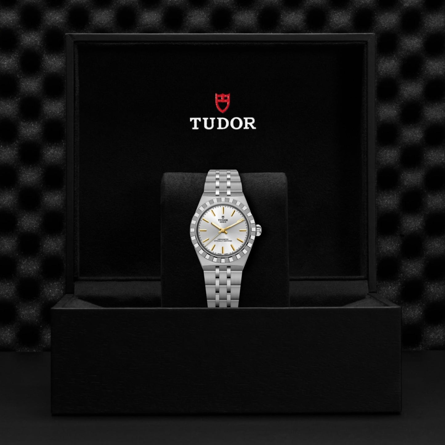 A TUDOR Royal wristwatch with a silver and gold face and metal bracelet is displayed upright in a black cushioned box, with the TUDOR logo and name visible on the inner lid.