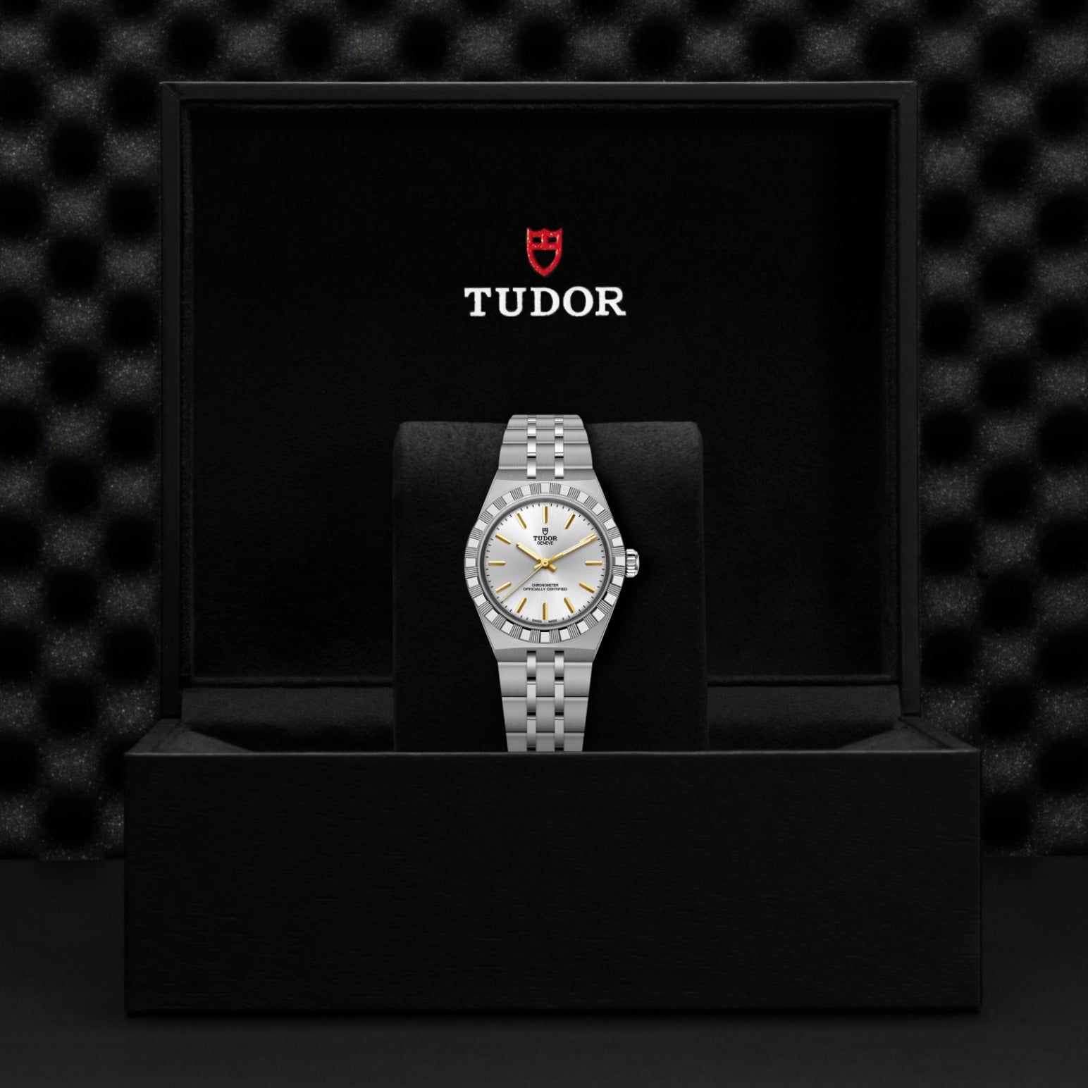 A TUDOR Royal wristwatch with a silver and gold face and metal bracelet is displayed upright in a black cushioned box, with the TUDOR logo and name visible on the inner lid.