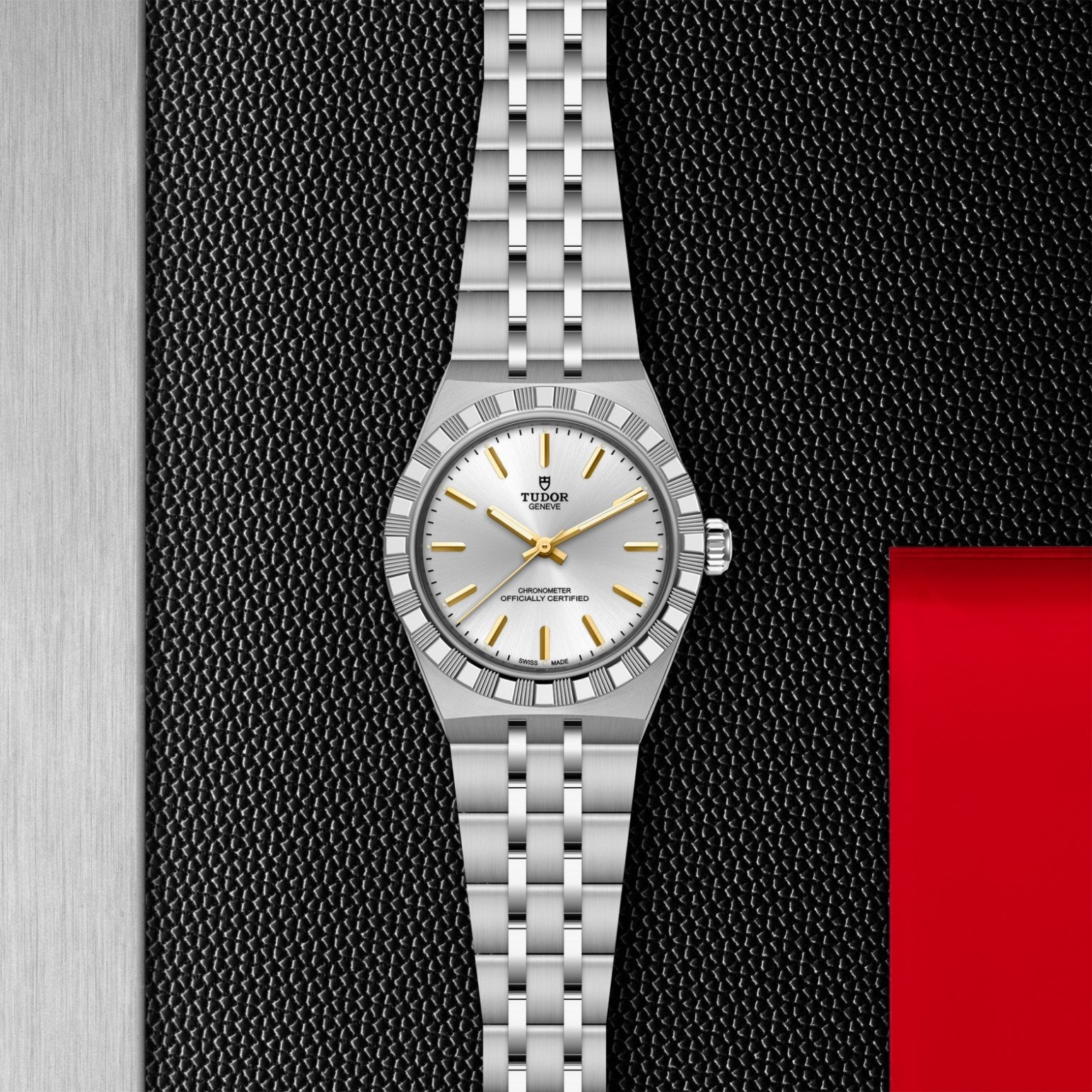 The TUDOR Royal by TUDOR features a silver case, white dial, gold hour markers, and a metal link bracelet. Displayed on a textured black surface with silver and red accents, it highlights the sport-chic elegance of automatic watches.