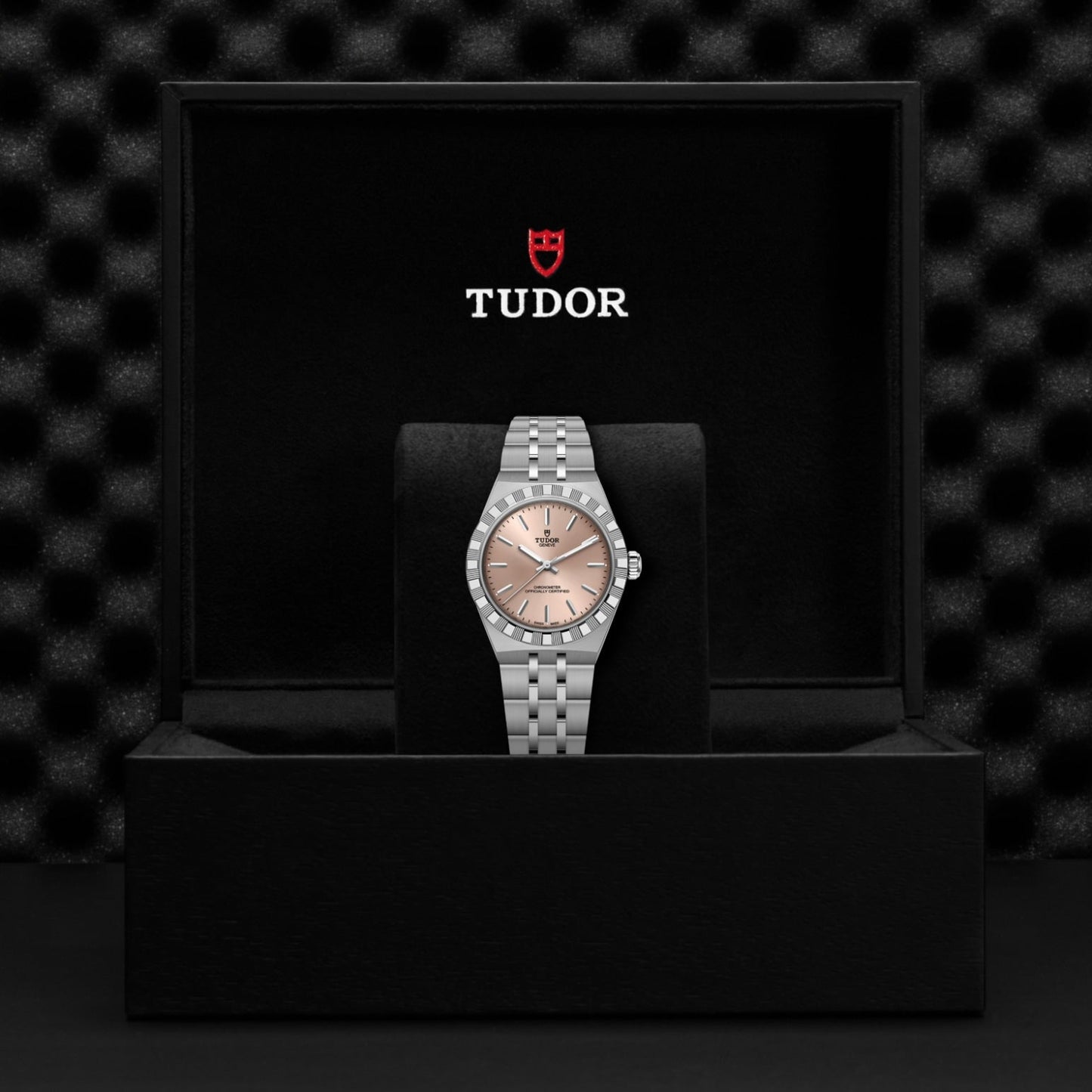 A TUDOR Royal wristwatch with a pink dial sits on a black cushion inside a box displaying the TUDOR logo and shield. The automatic sport-chic watch stands out against the textured black background.