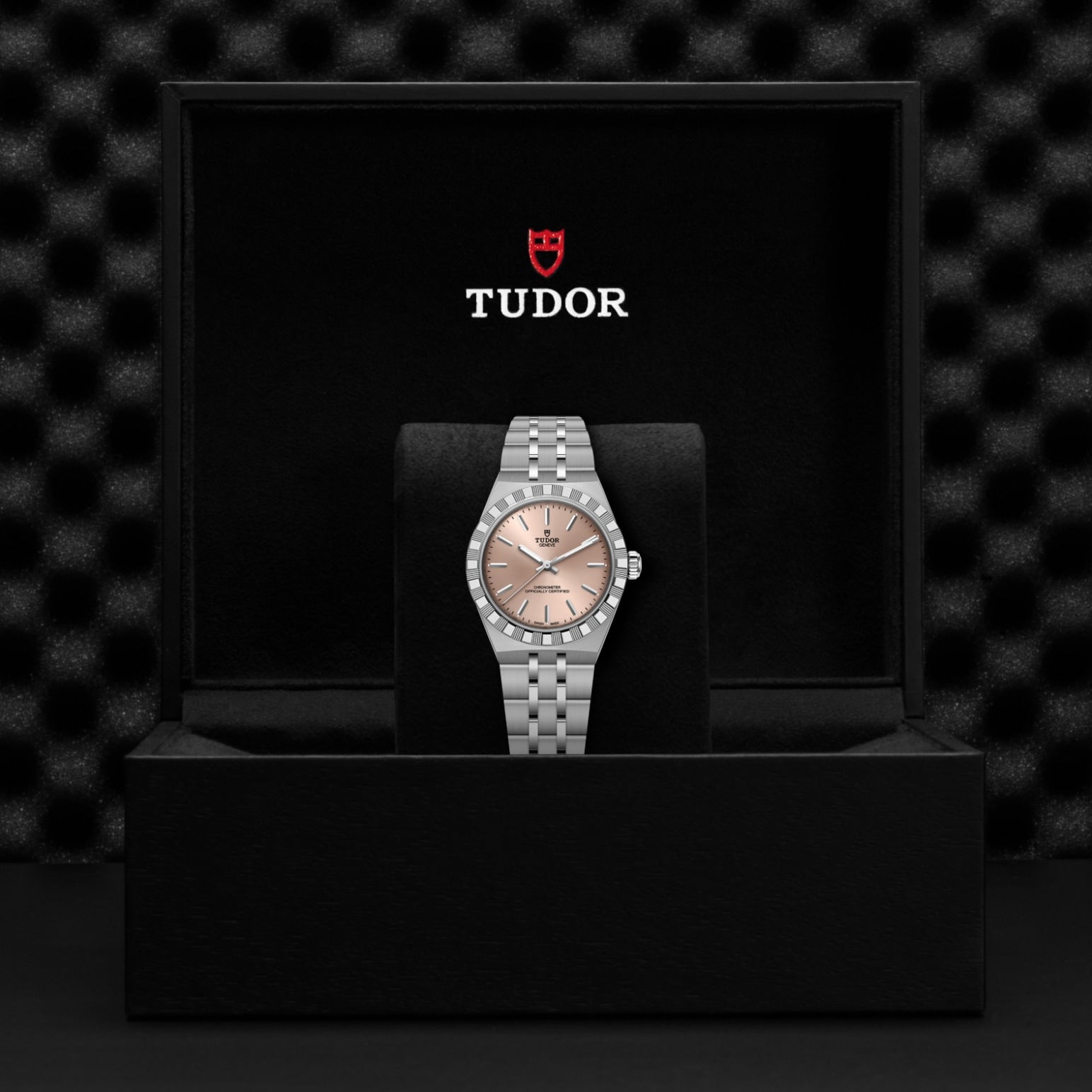 A TUDOR Royal wristwatch with a pink dial sits on a black cushion inside a box displaying the TUDOR logo and shield. The automatic sport-chic watch stands out against the textured black background.