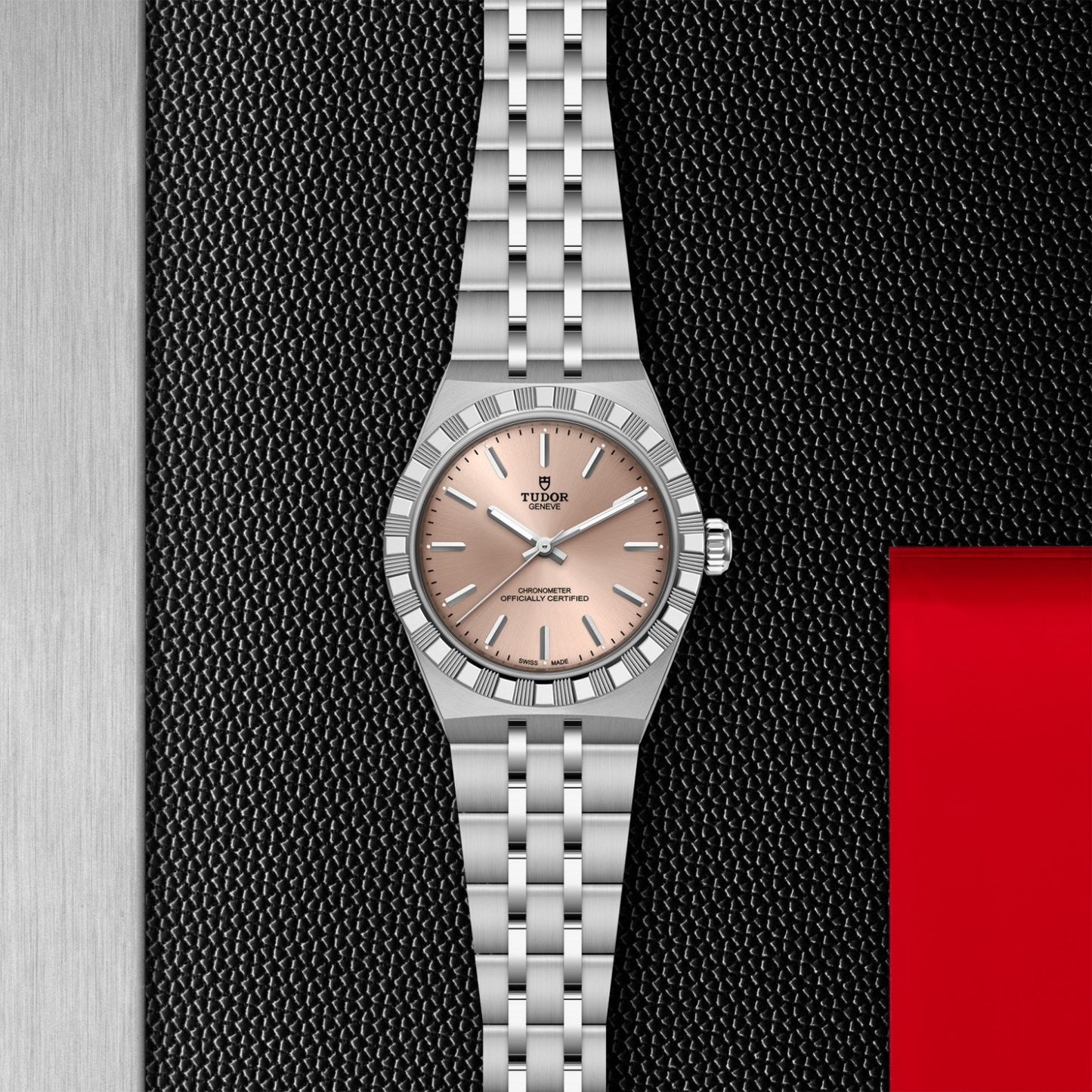 A TUDOR Royal watch by TUDOR, featuring a stainless steel bracelet and light pink dial, rests on a black textured surface with a red rectangle in the bottom right corner.
