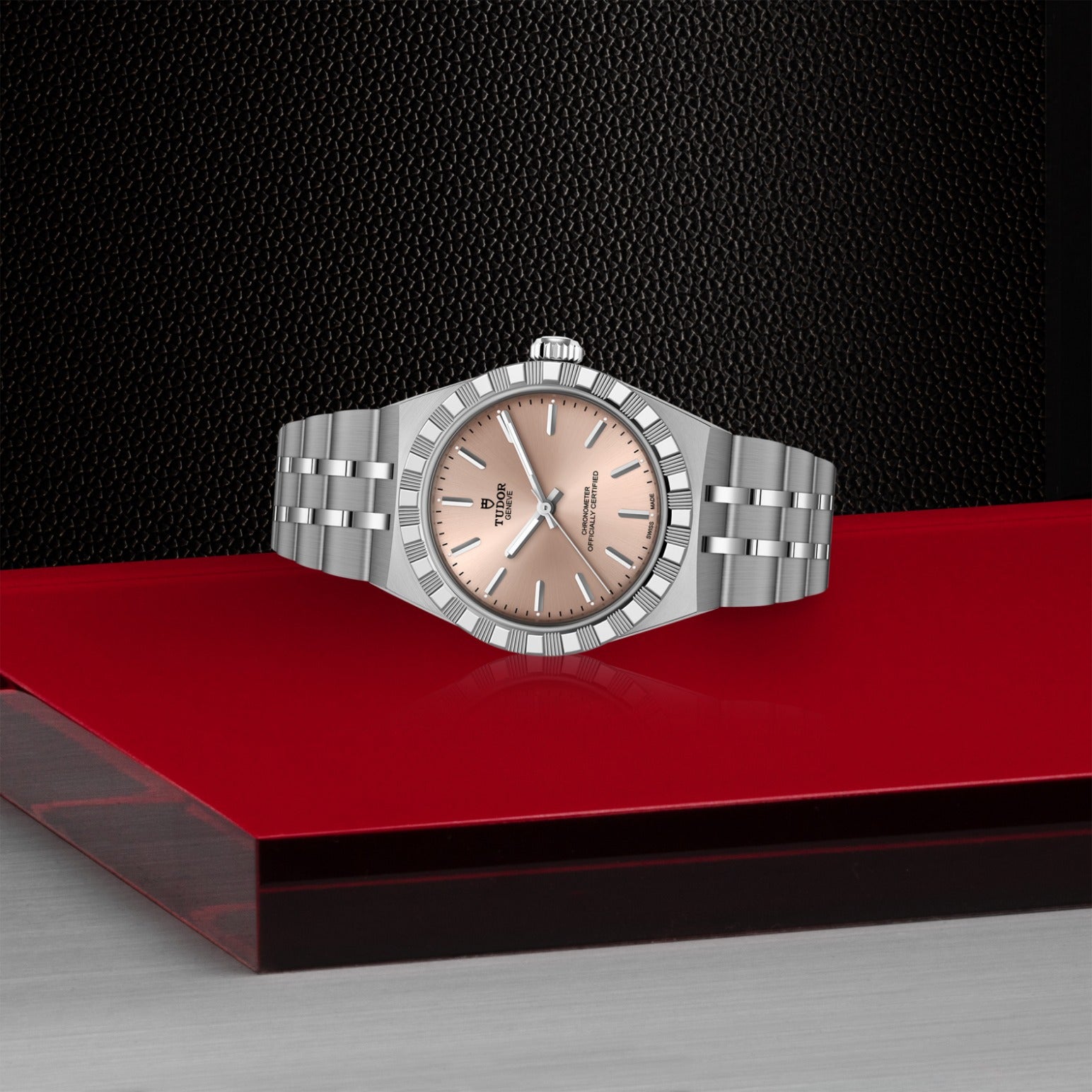 The TUDOR Royal by TUDOR features a silver case and rose gold dial, displayed on a red and silver geometric surface that highlights the watch's elegant stainless steel design against a dark textured background.