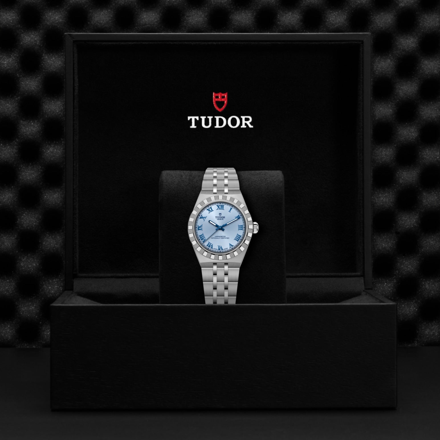 A TUDOR Royal stainless steel watch with a light blue dial rests on a black cushion inside an open TUDOR box, displaying the red Tudor logo and white branding text on the inner lid.