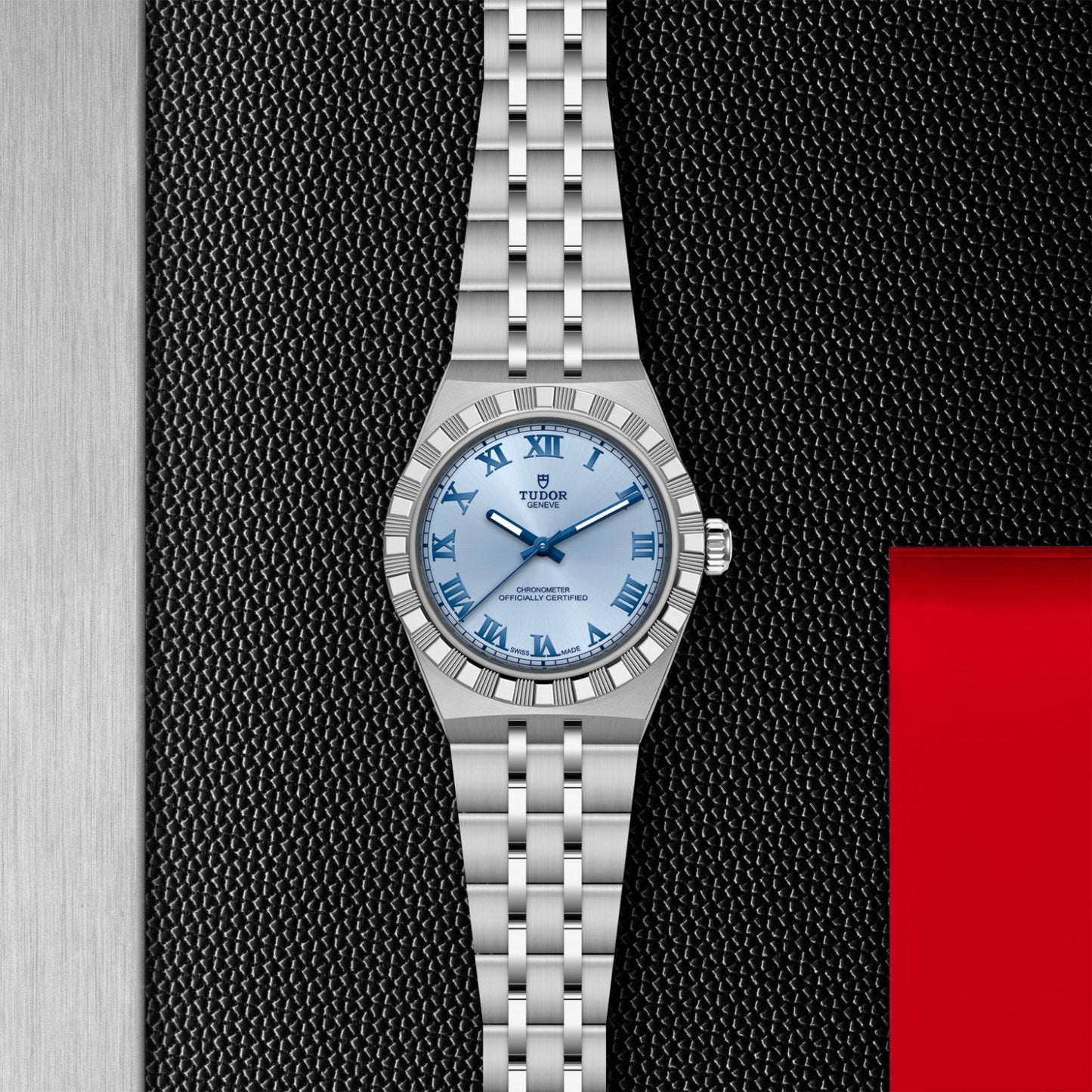 The TUDOR Royal by TUDOR features a silver metal band and light blue dial with silver Roman numerals, presented on a textured black surface with metallic and red accents—highlighting its automatic sport-chic elegance.