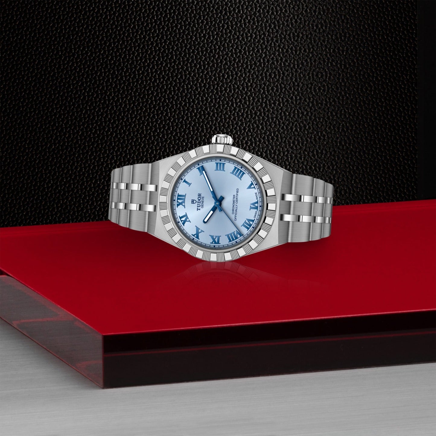 A TUDOR Royal wristwatch by TUDOR features a silver case and blue Roman numeral dial, resting on a glossy red surface with a black and gray backdrop, exemplifying the elegance of automatic sport-chic timepieces.