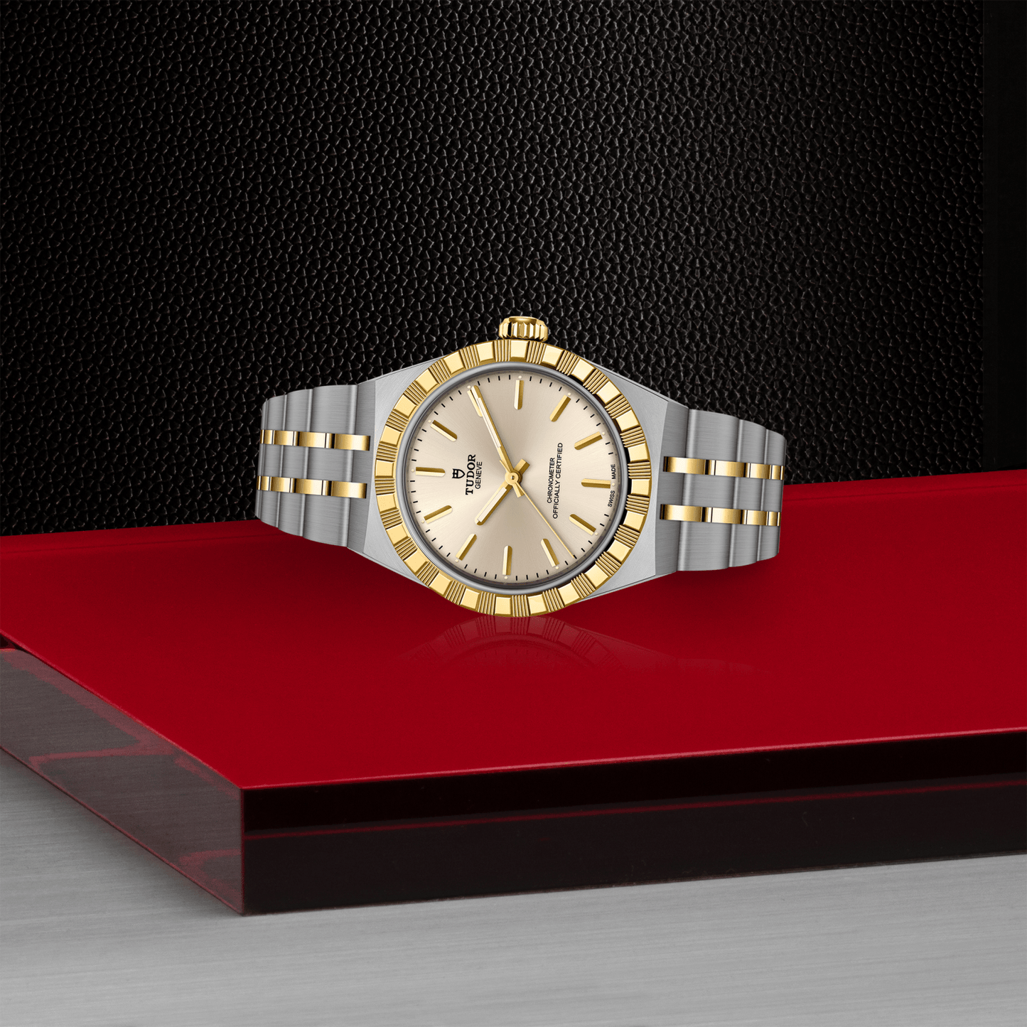 The TUDOR Royal wristwatch by TUDOR, featuring a stainless steel and gold band, gold bezel, and champagne dial, is showcased on a red and gray surface with a black textured background.