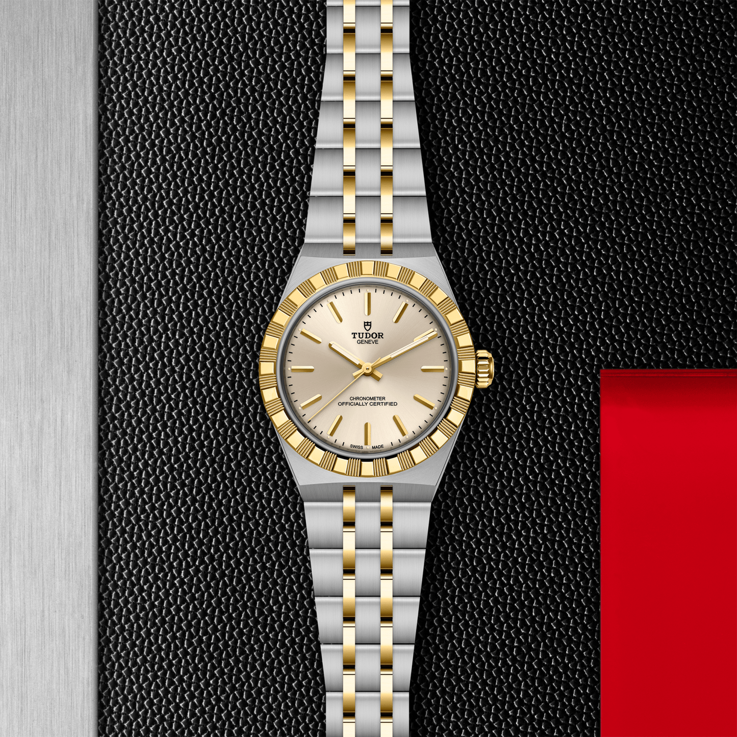 The TUDOR Royal by TUDOR features a stainless steel and gold bracelet, a gold bezel, and a silver dial with gold markers and hands, displayed on a textured black background with metallic and red accents.