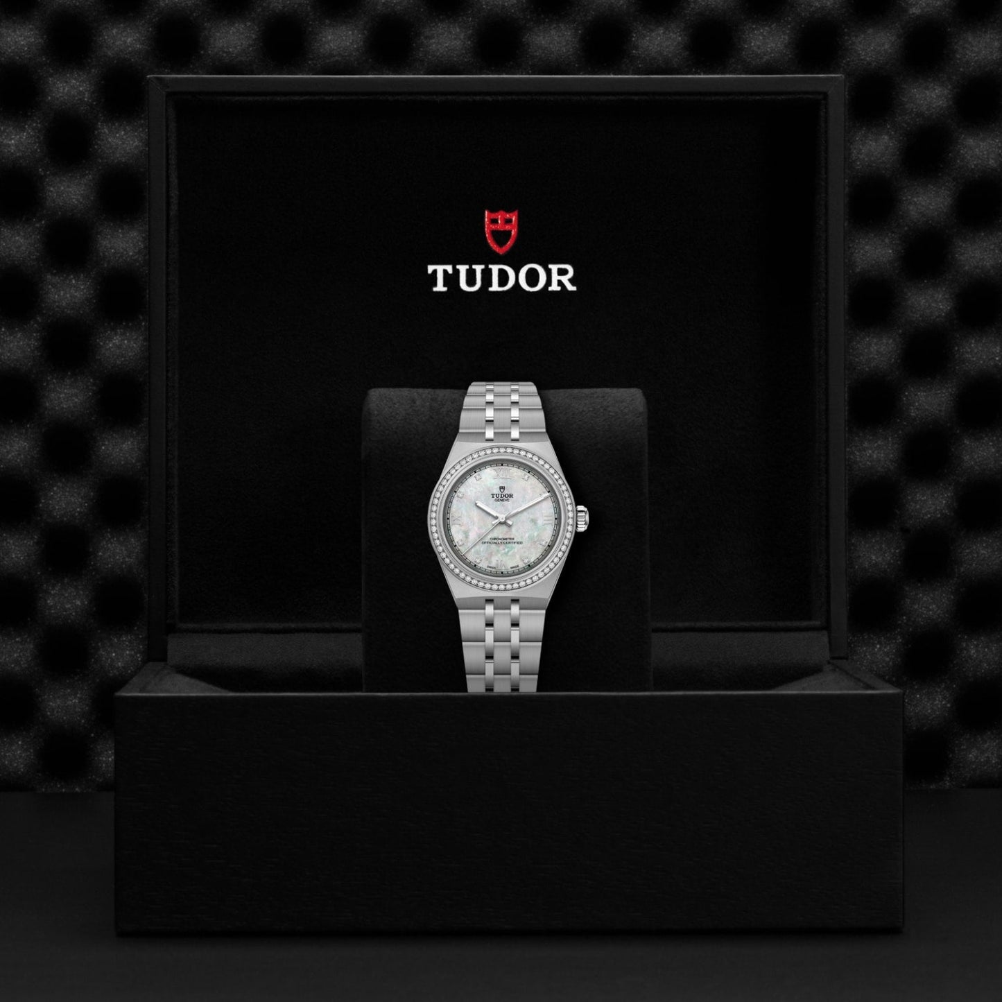 A TUDOR Royal wristwatch with a diamond bezel and mother-of-pearl dial, featuring an integrated bracelet, is displayed on a black cushion inside an open TUDOR box with the logo and brand name on the inside lid.