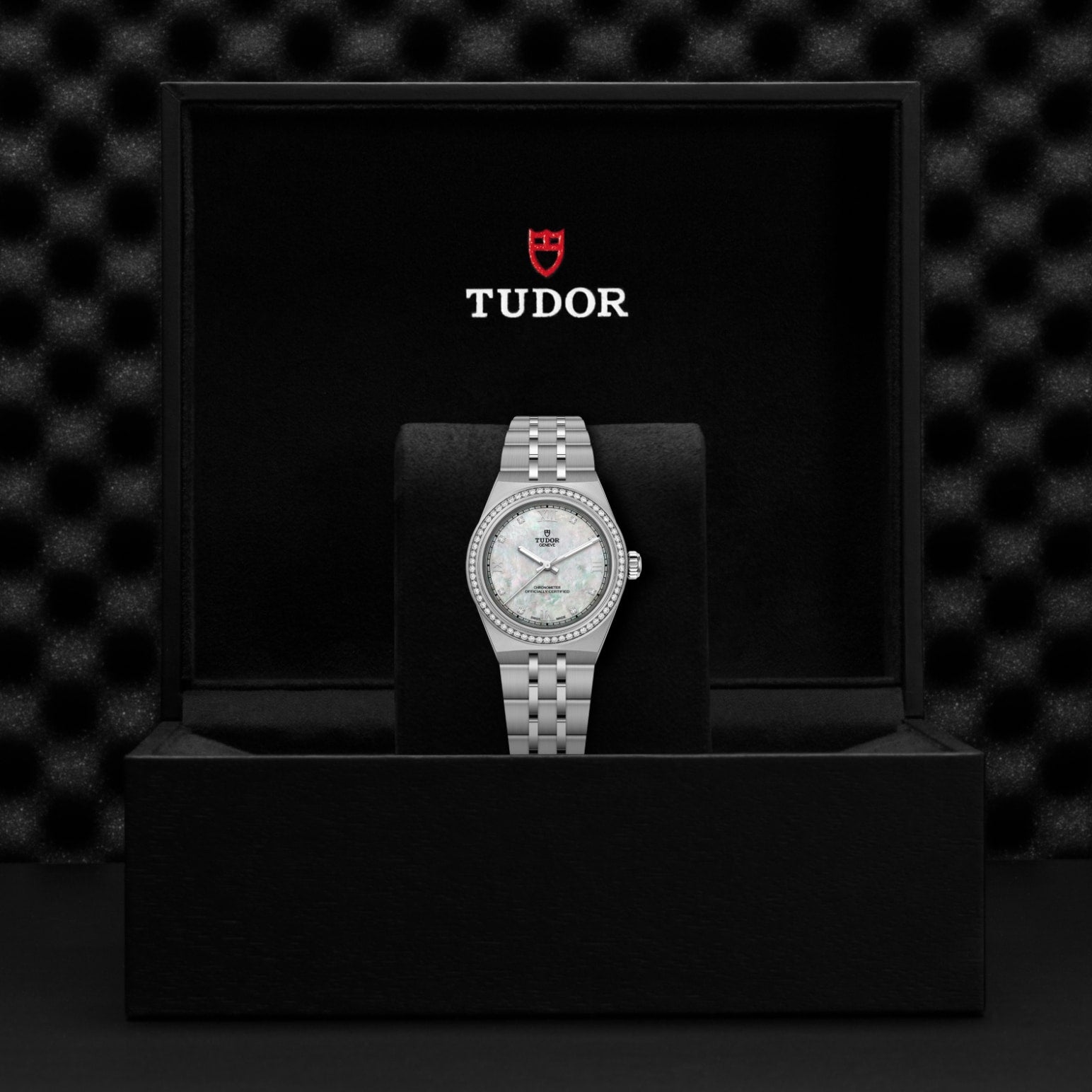 A TUDOR Royal wristwatch with a diamond bezel and mother-of-pearl dial, featuring an integrated bracelet, is displayed on a black cushion inside an open TUDOR box with the logo and brand name on the inside lid.
