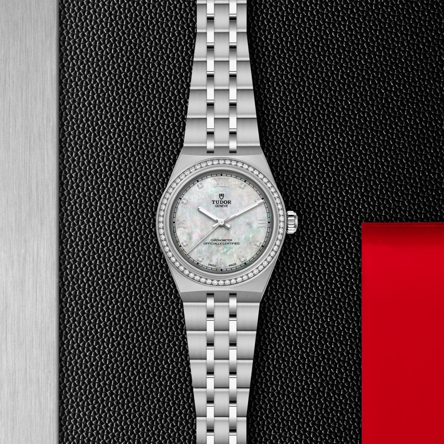 The TUDOR Royal wristwatch by TUDOR features a silver case, white mother-of-pearl dial with diamond markers, and integrated bracelet, displayed on a textured black and metallic background with a red accent at the bottom right.