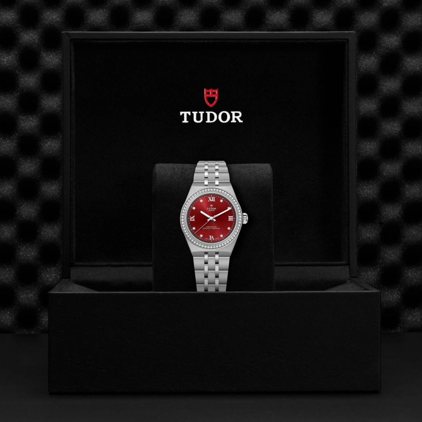 A silver TUDOR Royal wristwatch with a red dial and diamond markers stands upright in a black box featuring "TUDOR" and its shield logo in white and red on the lid, exemplifying the elegance of TUDOR Royal's automatic sport-chic style.