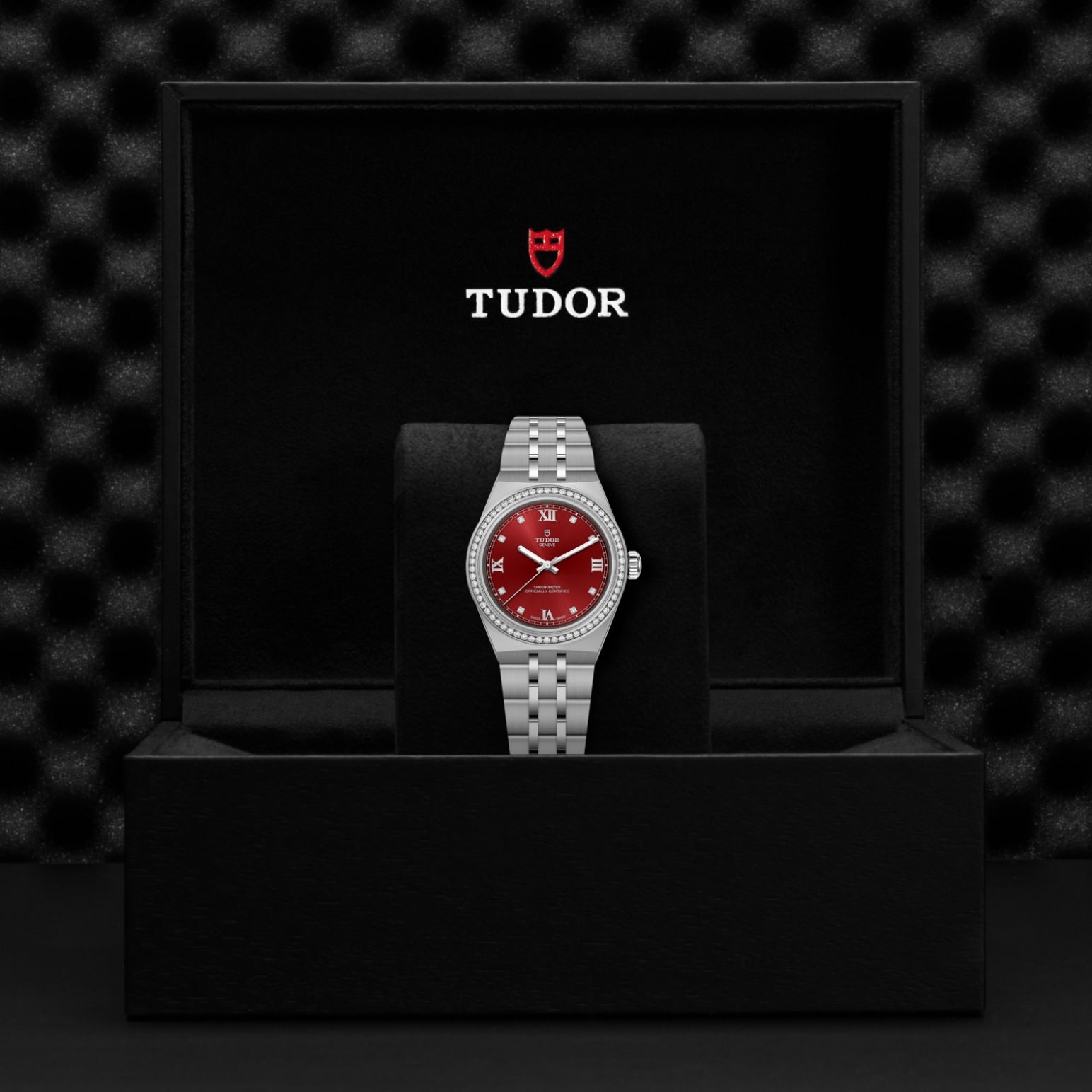 A silver TUDOR Royal wristwatch with a red dial and diamond markers stands upright in a black box featuring "TUDOR" and its shield logo in white and red on the lid, exemplifying the elegance of TUDOR Royal's automatic sport-chic style.