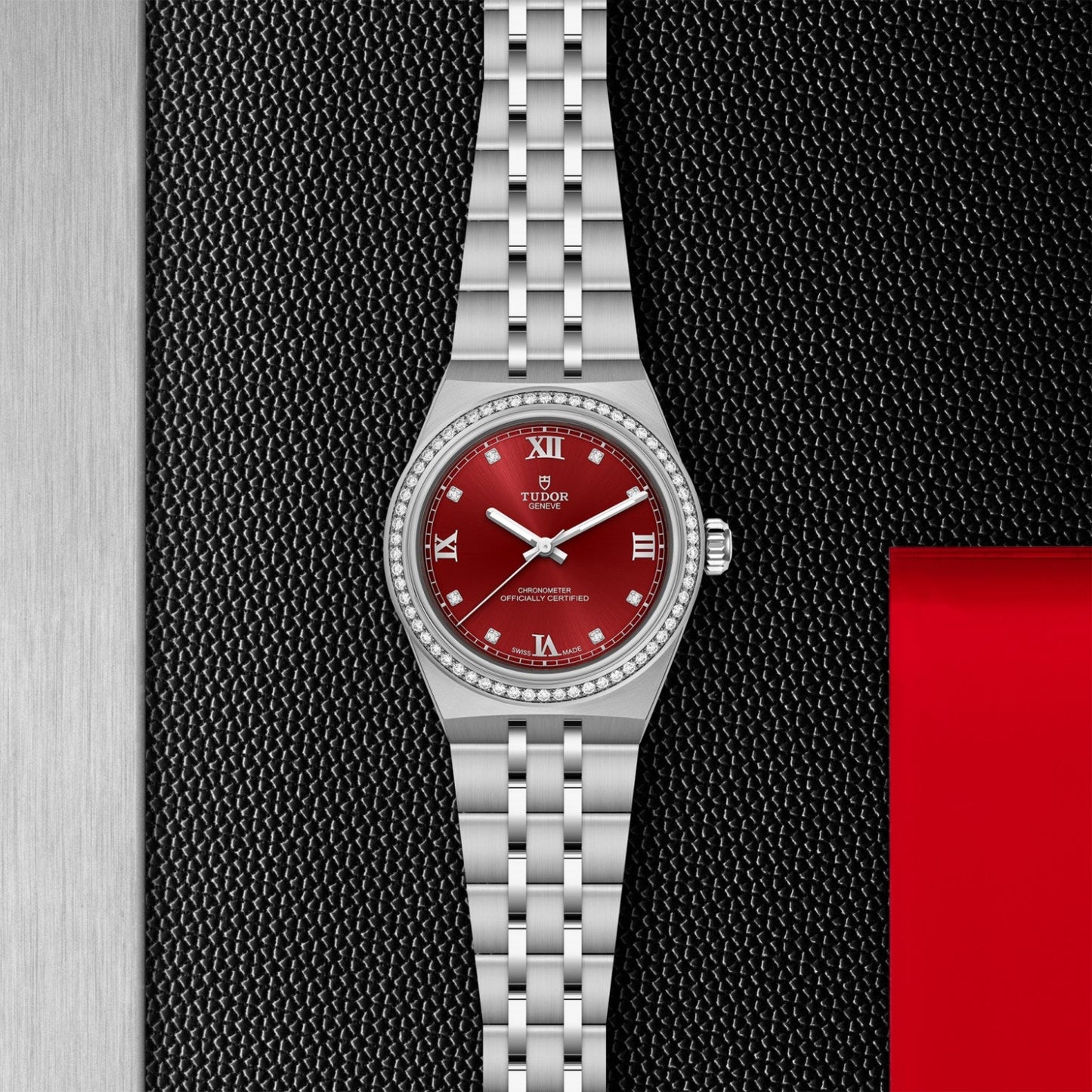 The TUDOR Royal by TUDOR features a stainless steel case, red dial, silver hour markers, and date window, showcased on a black and silver textured background with a red accent on the right.