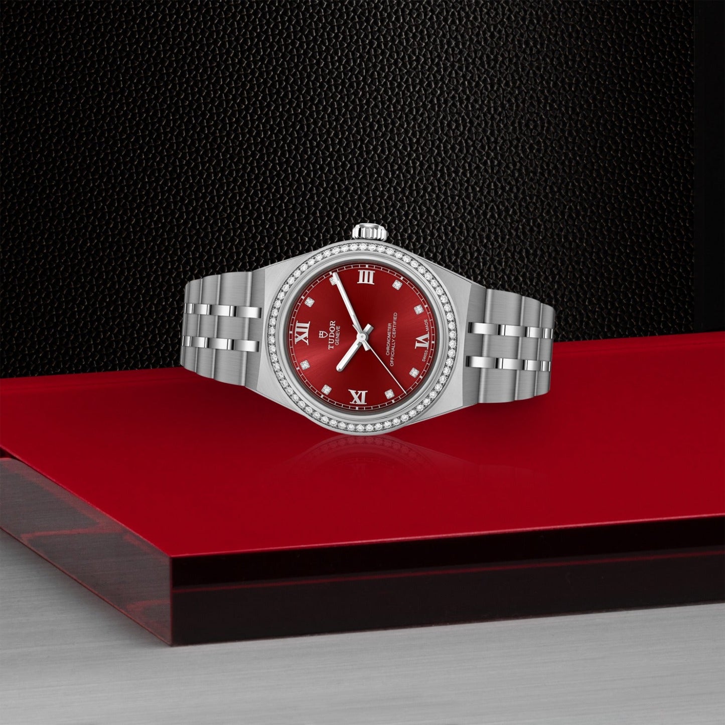 The TUDOR Royal by TUDOR, a silver luxury wristwatch with a red dial and Roman numerals, is showcased on a glossy red surface against a textured black background, capturing the allure of automatic sport-chic design.