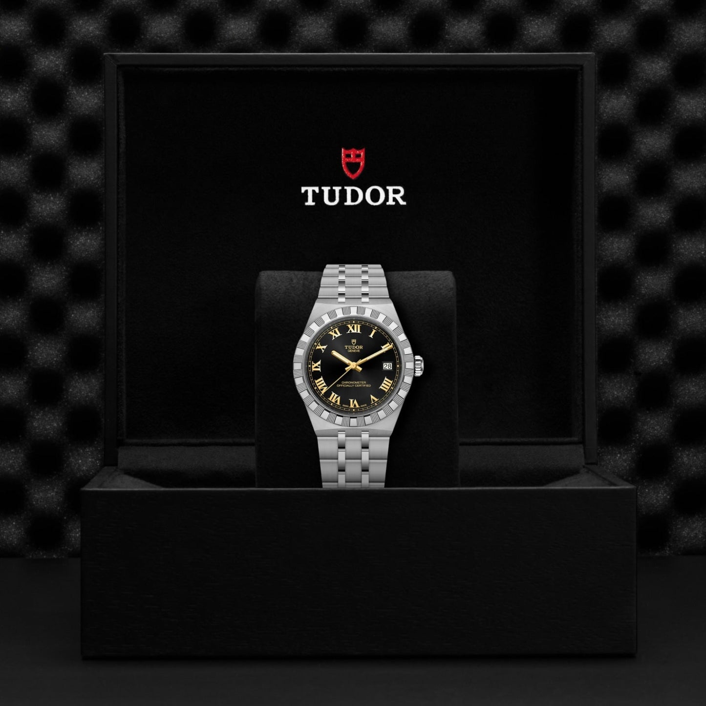 A TUDOR Royal wristwatch by TUDOR, featuring a black and gold dial, stands upright in an open black box with the Tudor logo inside the lid. A textured black background highlights this refined automatic sport-chic timepiece.
