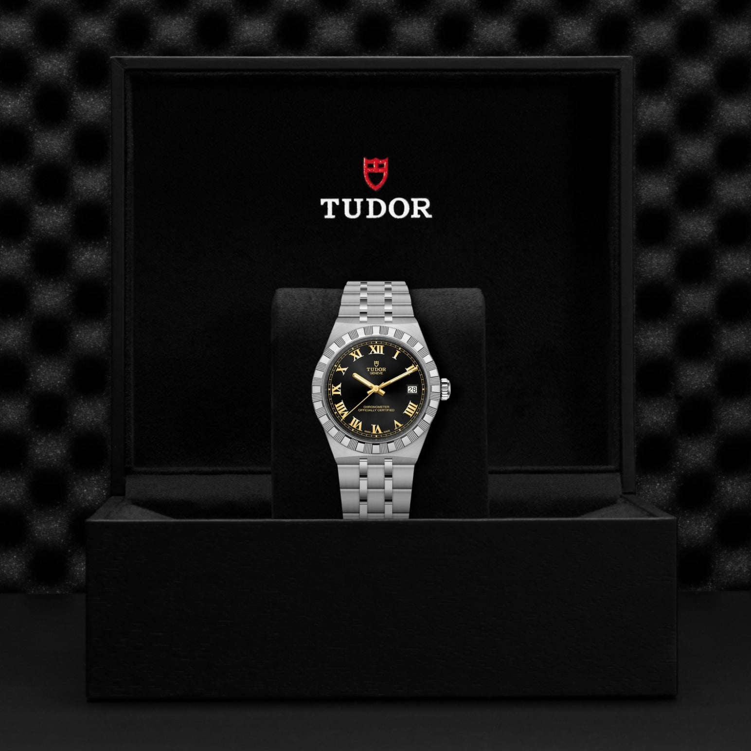A TUDOR Royal wristwatch by TUDOR, featuring a black and gold dial, stands upright in an open black box with the Tudor logo inside the lid. A textured black background highlights this refined automatic sport-chic timepiece.