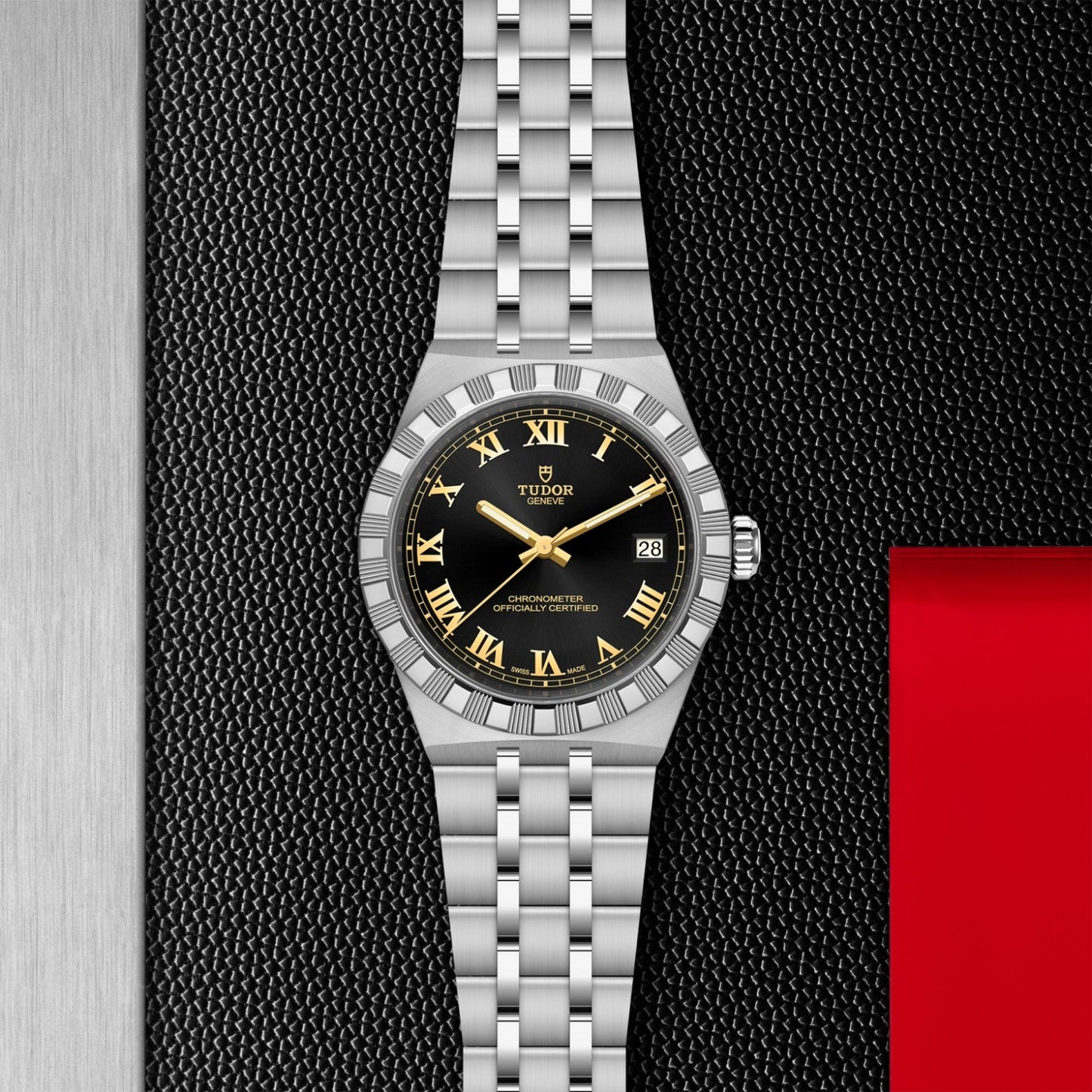 A TUDOR Royal wristwatch by TUDOR featuring a silver stainless steel bracelet, black dial, and gold Roman numerals rests on a textured black surface with metal details and red accents around it.