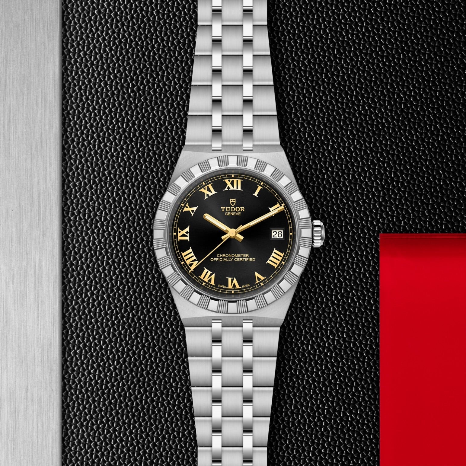 A TUDOR Royal wristwatch by TUDOR featuring a silver stainless steel bracelet, black dial, and gold Roman numerals rests on a textured black surface with metal details and red accents around it.