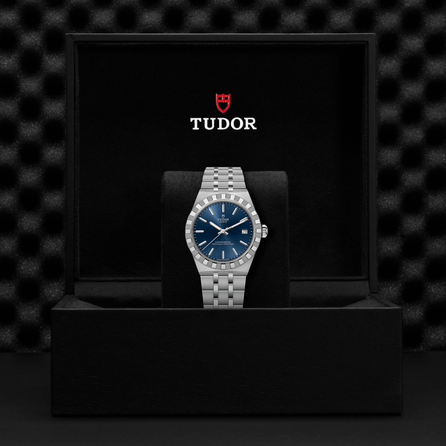 A TUDOR Royal wristwatch is shown in an open black TUDOR box against a dark textured background, featuring a blue dial and stainless steel case.