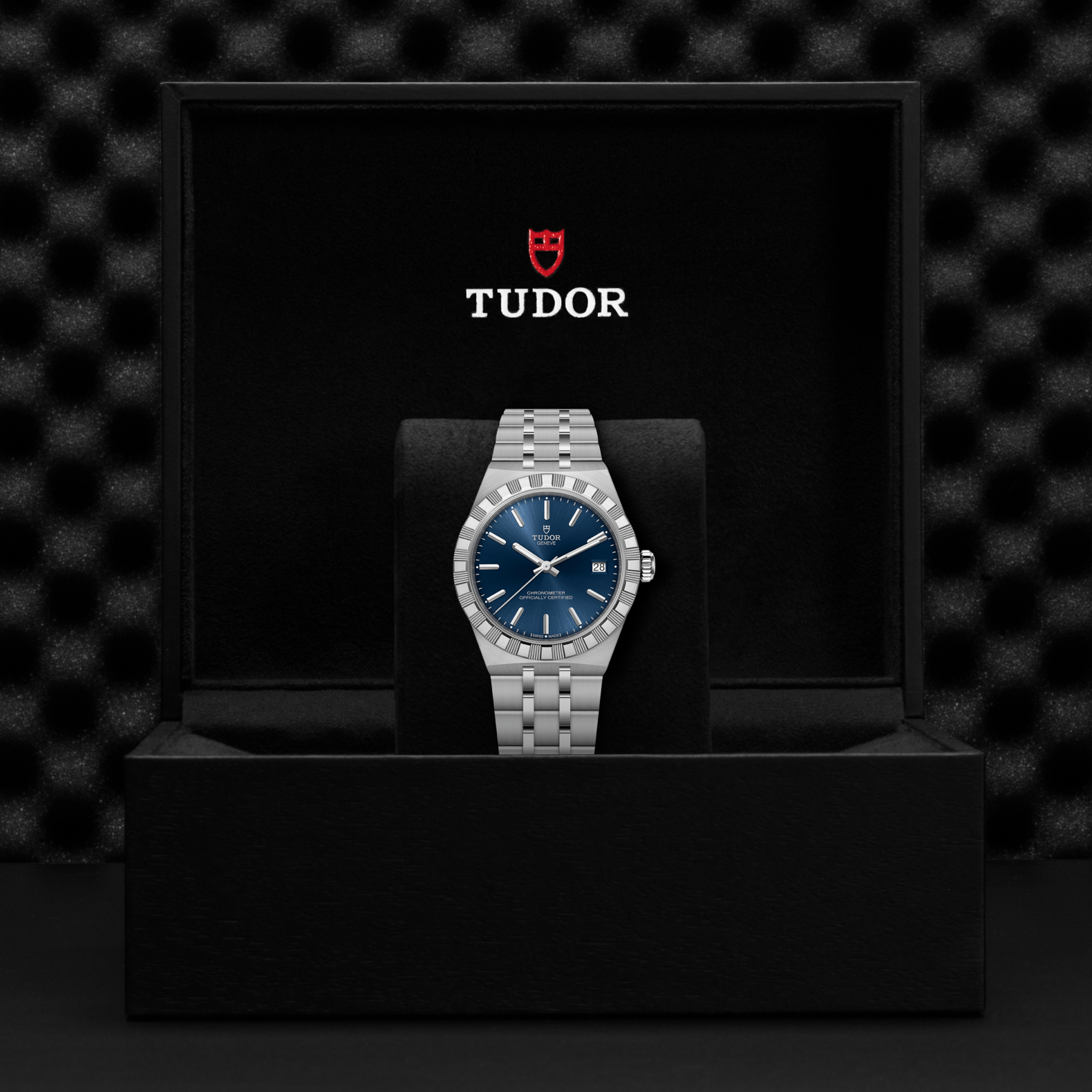 A TUDOR Royal wristwatch is shown in an open black TUDOR box against a dark textured background, featuring a blue dial and stainless steel case.