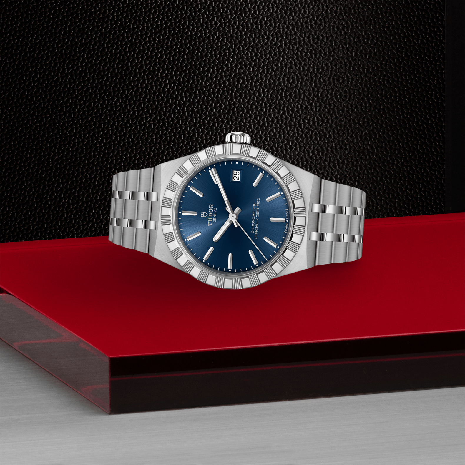 A TUDOR Royal wristwatch by TUDOR, featuring a blue dial, date display, and stainless steel case, is placed on a red and black geometric surface with a dark textured background.