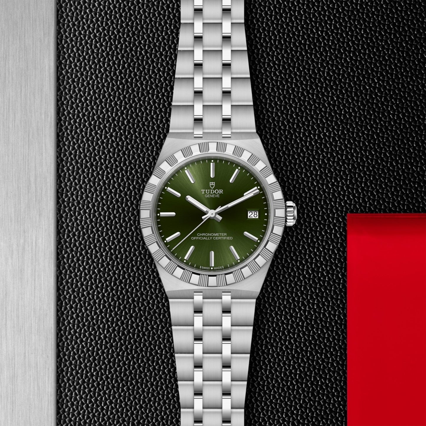 The TUDOR Royal by TUDOR, a stainless steel watch with green dial, date display, and metal bracelet, is set on a textured black surface with metallic and red accents.