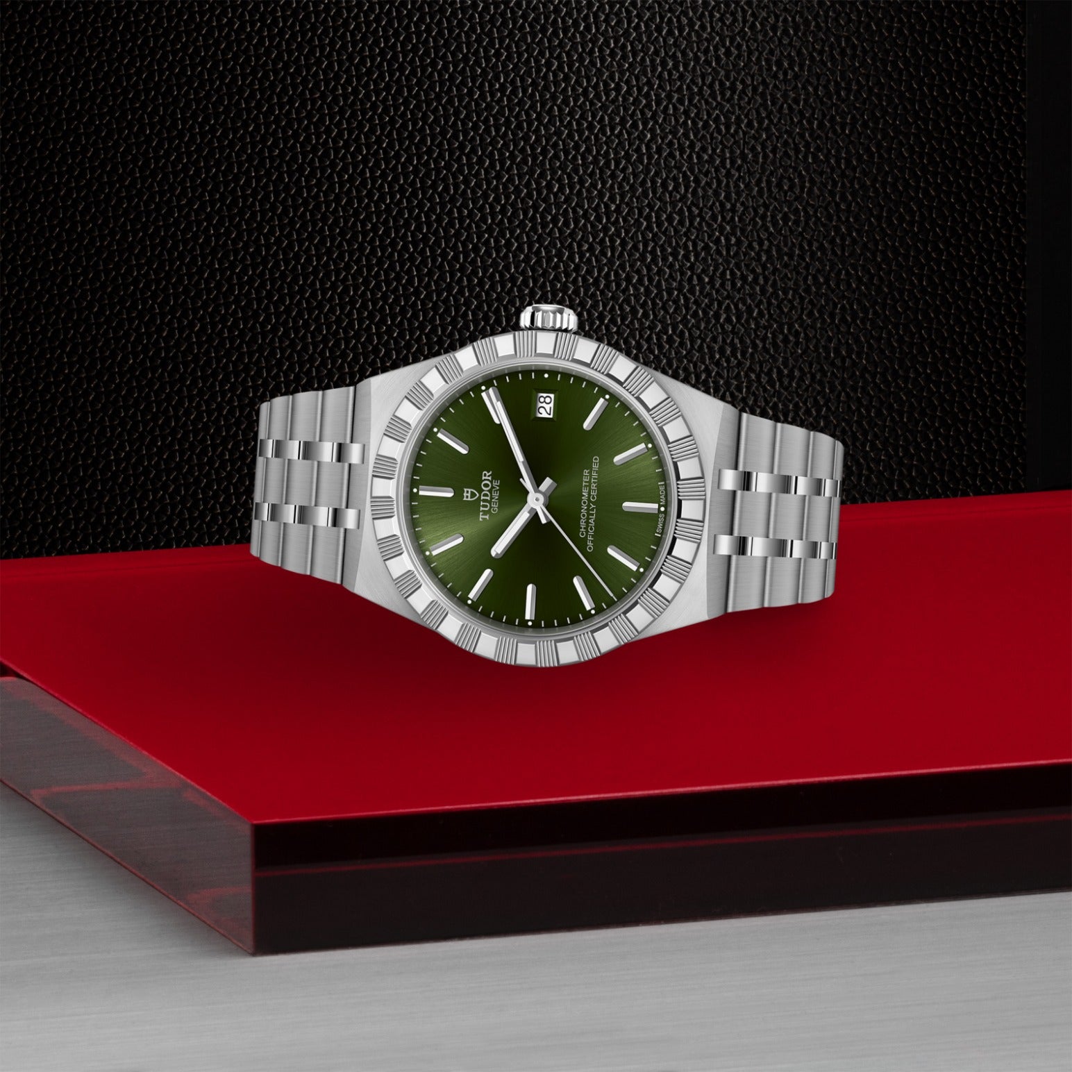 A TUDOR Royal wristwatch by TUDOR, featuring a metallic bracelet and green dial with silver markers, hands, and date display, is showcased on a red and gray platform against a textured black background.