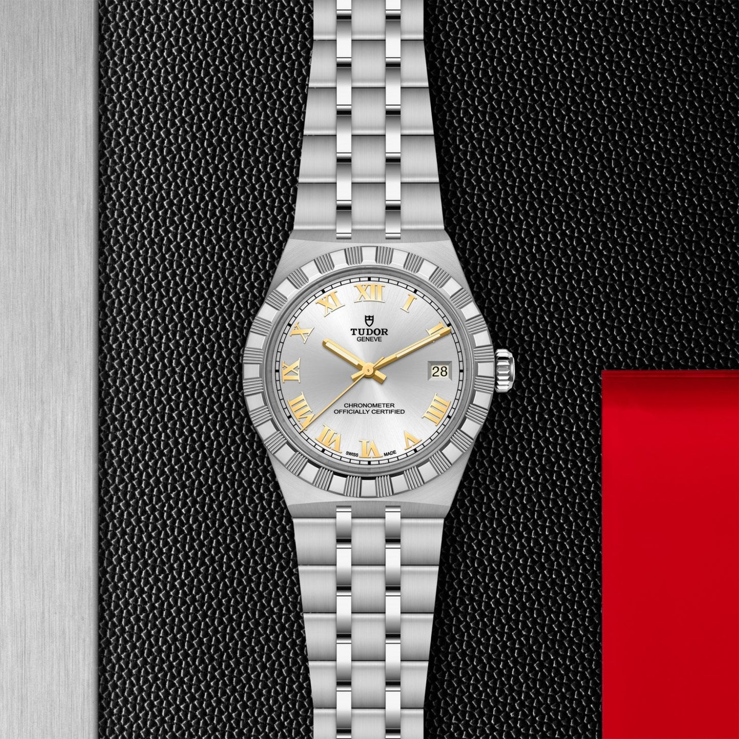 The TUDOR Royal by TUDOR features a silver case, white dial, gold hour markers, and a date display at 3 o'clock—an automatic sport-chic wristwatch ideal for enthusiasts. Shown on a textured black surface with metallic and red accents.