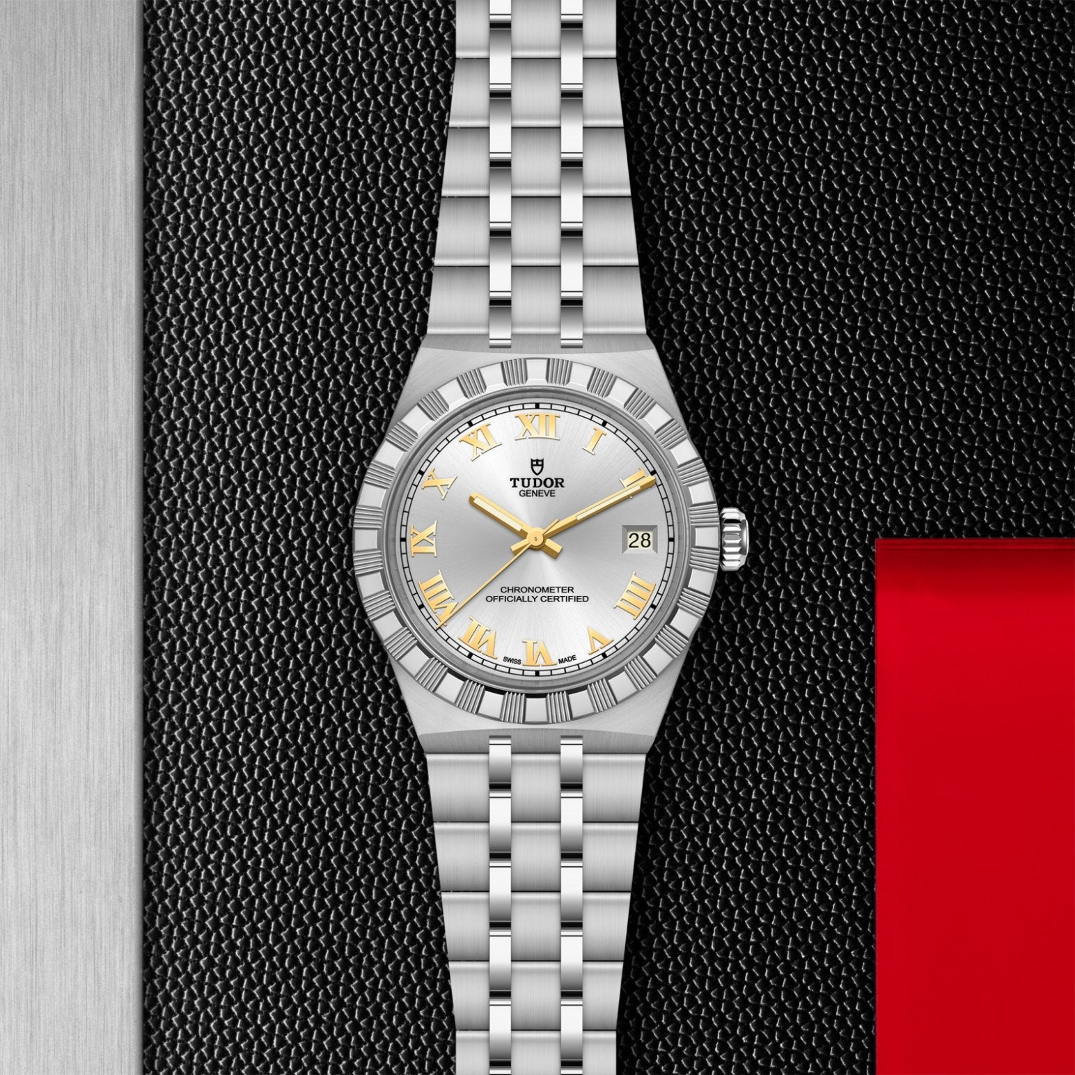The TUDOR Royal by TUDOR features a silver case, white dial, gold hour markers, and a date display at 3 o'clock—an automatic sport-chic wristwatch ideal for enthusiasts. Shown on a textured black surface with metallic and red accents.