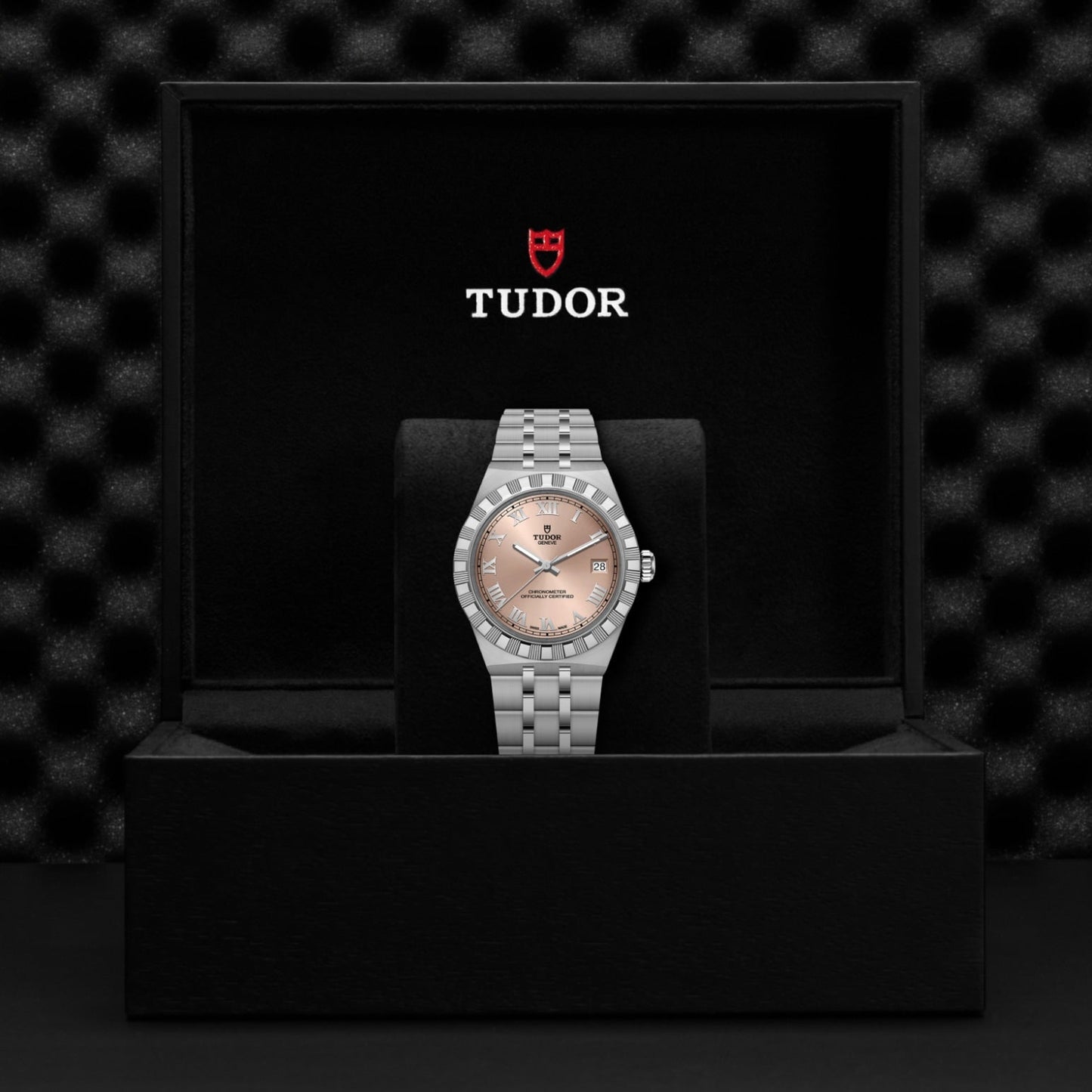 A silver TUDOR Royal wristwatch with a pink dial and date display is displayed on a cushion in a black TUDOR box against a textured black backdrop—an elegant presentation of the TUDOR Royal’s automatic sport-chic design.