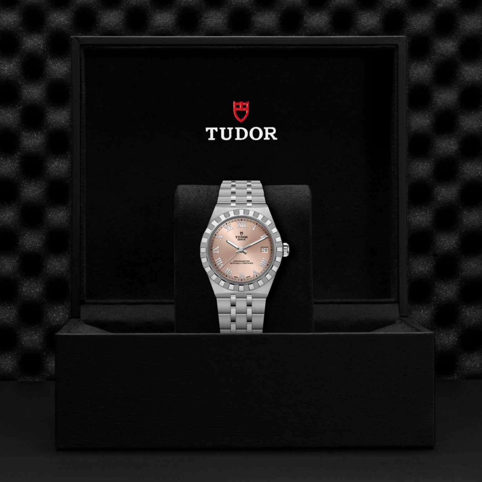 A silver TUDOR Royal wristwatch with a pink dial and date display is displayed on a cushion in a black TUDOR box against a textured black backdrop—an elegant presentation of the TUDOR Royal’s automatic sport-chic design.