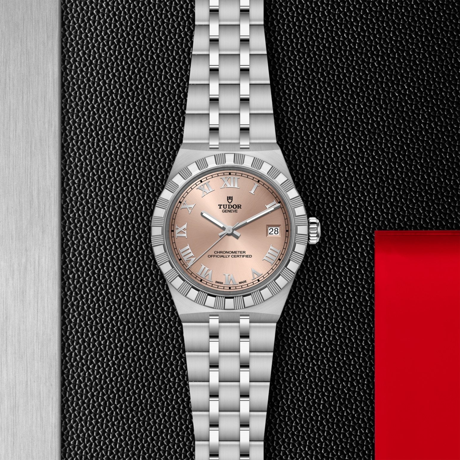 A TUDOR Royal wristwatch by TUDOR, featuring a salmon pink dial, Roman numeral bezel, and date display, rests on a textured black surface with metallic silver edges and a red accent.